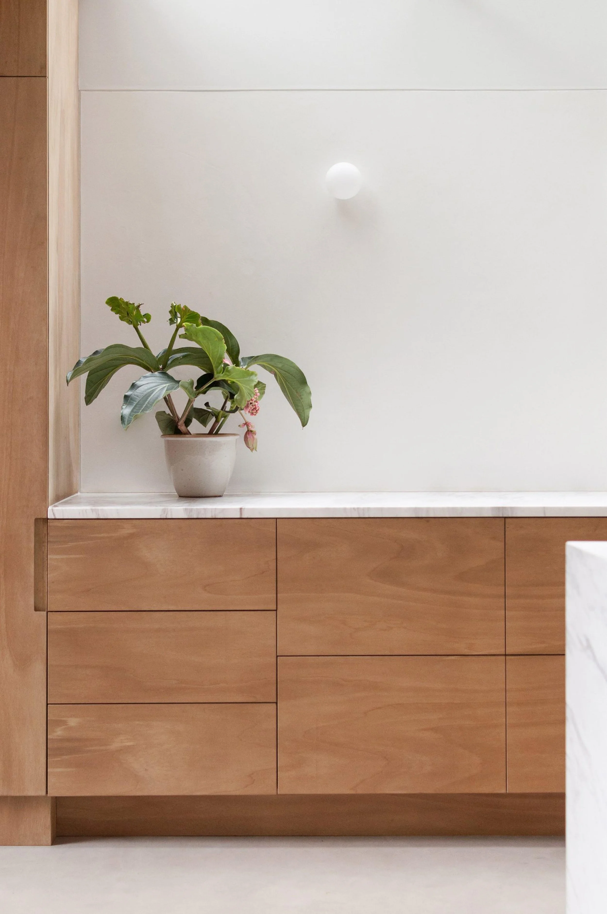 A minimalist interior with light wood cabinets, a white marble countertop, a potted green plant, and a white wall with a spherical wall sconce.
