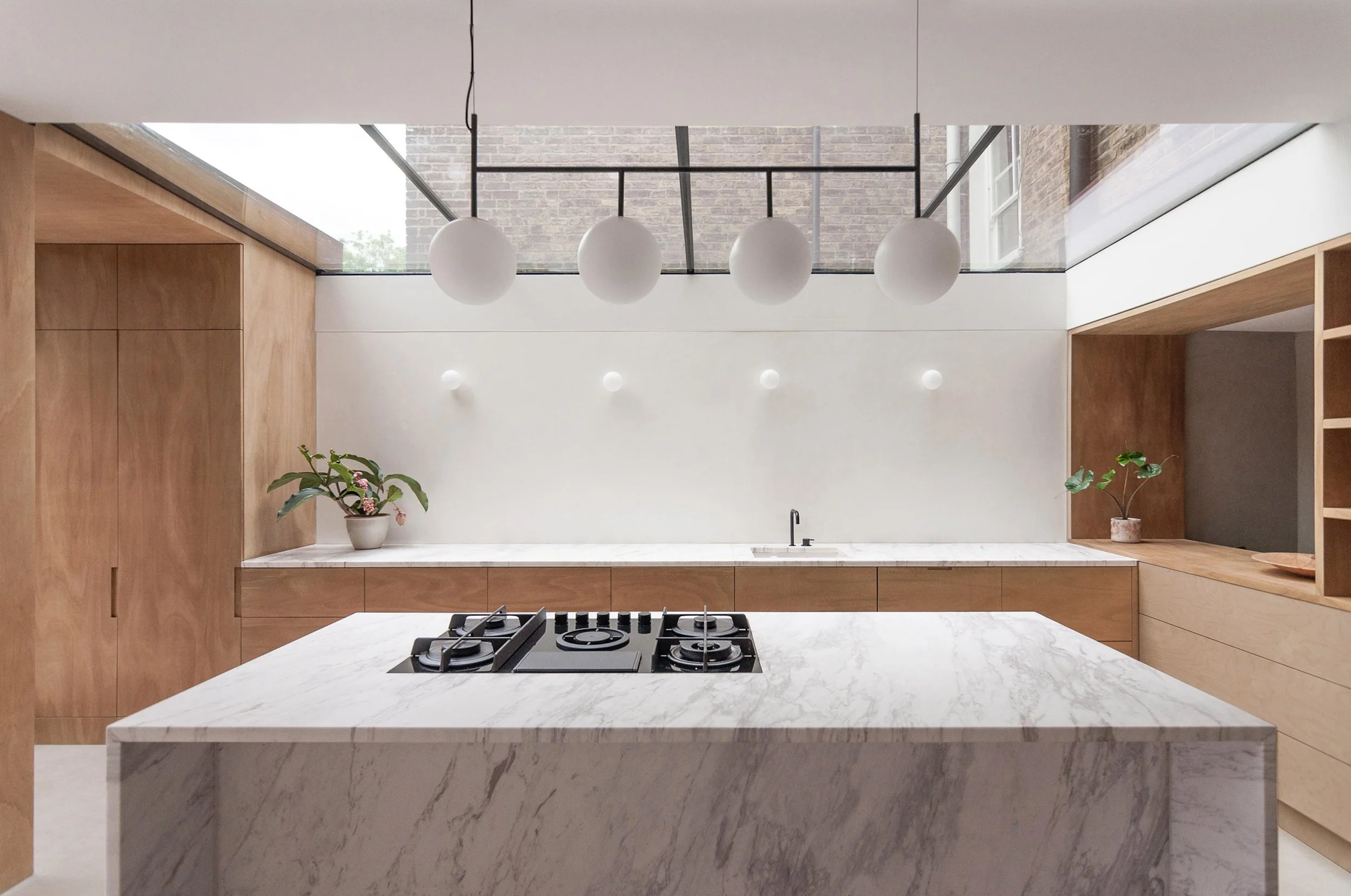 Modern kitchen with white marble island, wooden cabinets, and a white wall with small round wall lights. Four round pendant lights hang above the island, and there are potted plants on the countertops.