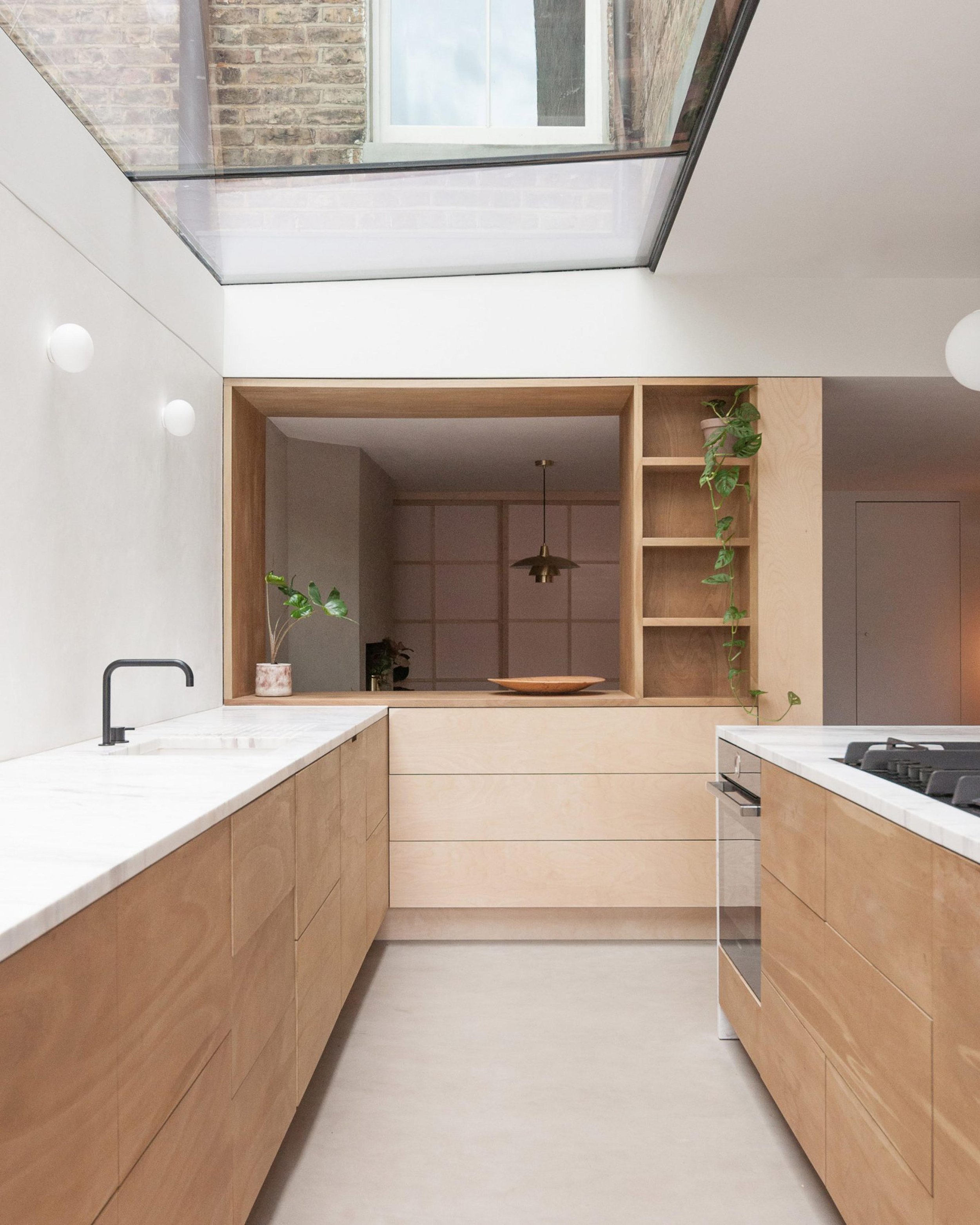 Modern kitchen with light wood cabinets, white marble countertops, black faucet, open shelving with plants, large skylight, and focusing on minimalist design.