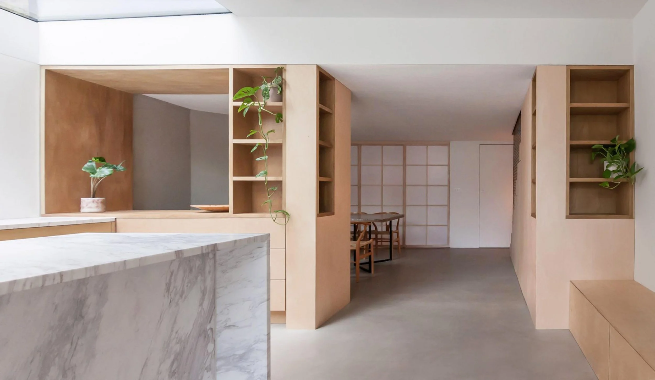 Minimalist interior with light wood shelving, potted plants, a marble countertop, and a dining area with wooden chairs and a table in the background.