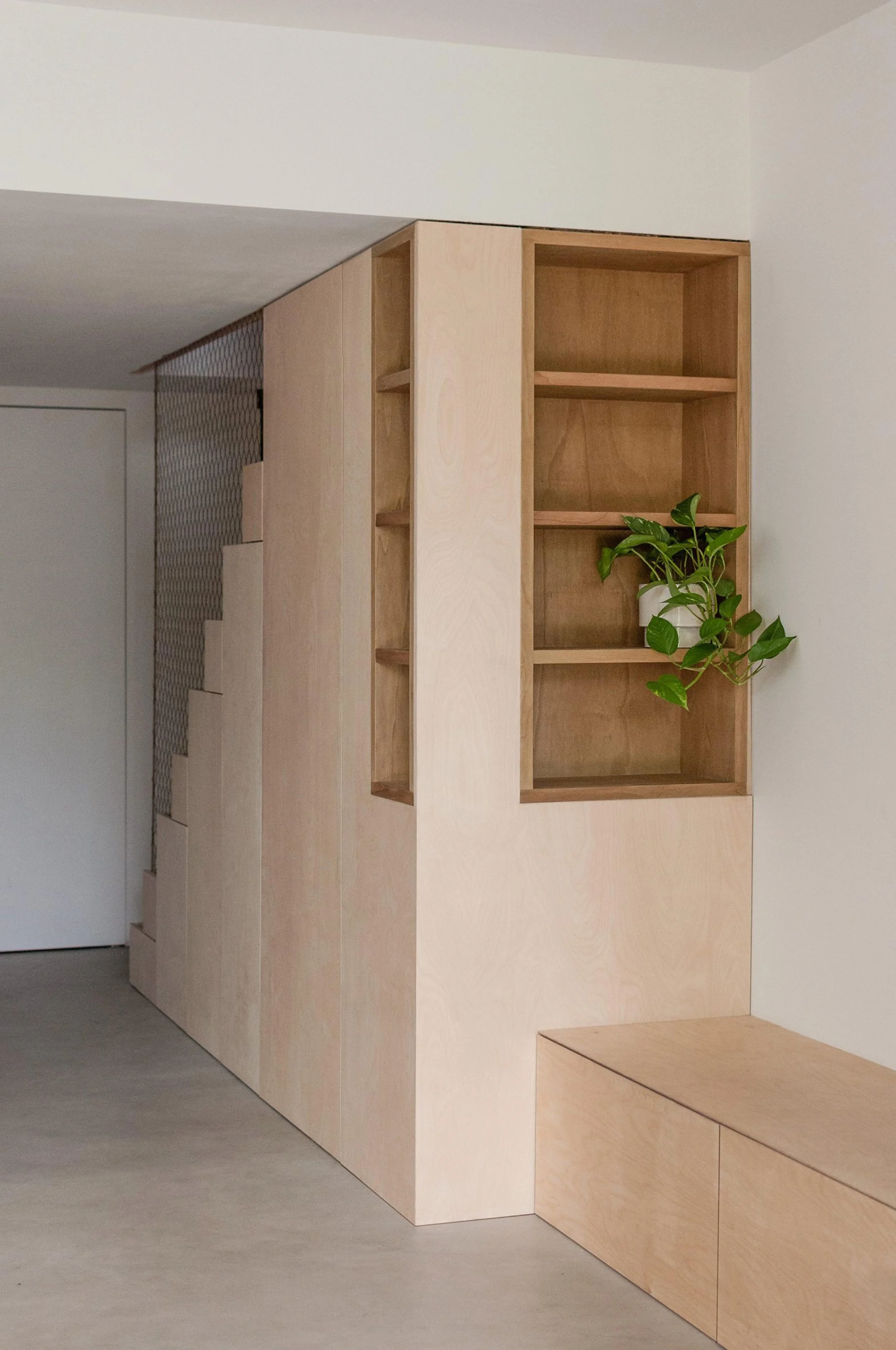 Interior view of a minimalist room featuring wooden shelving and a built-in storage bench, with a partially visible staircase and patterned wall in the background.