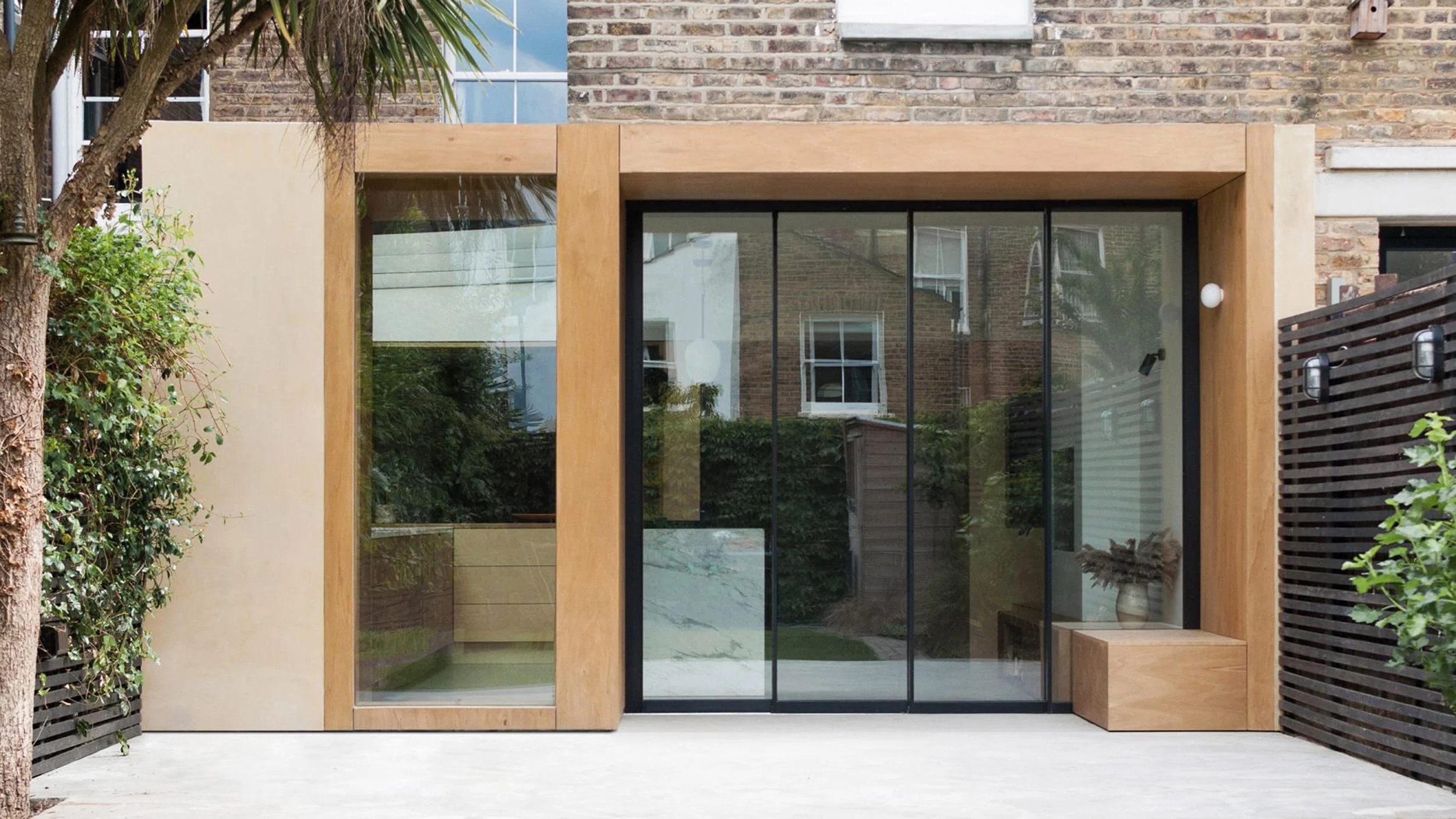 Modern house exterior with large glass sliding door framed in wood and beige walls, flanked by greenery and outdoor patio.