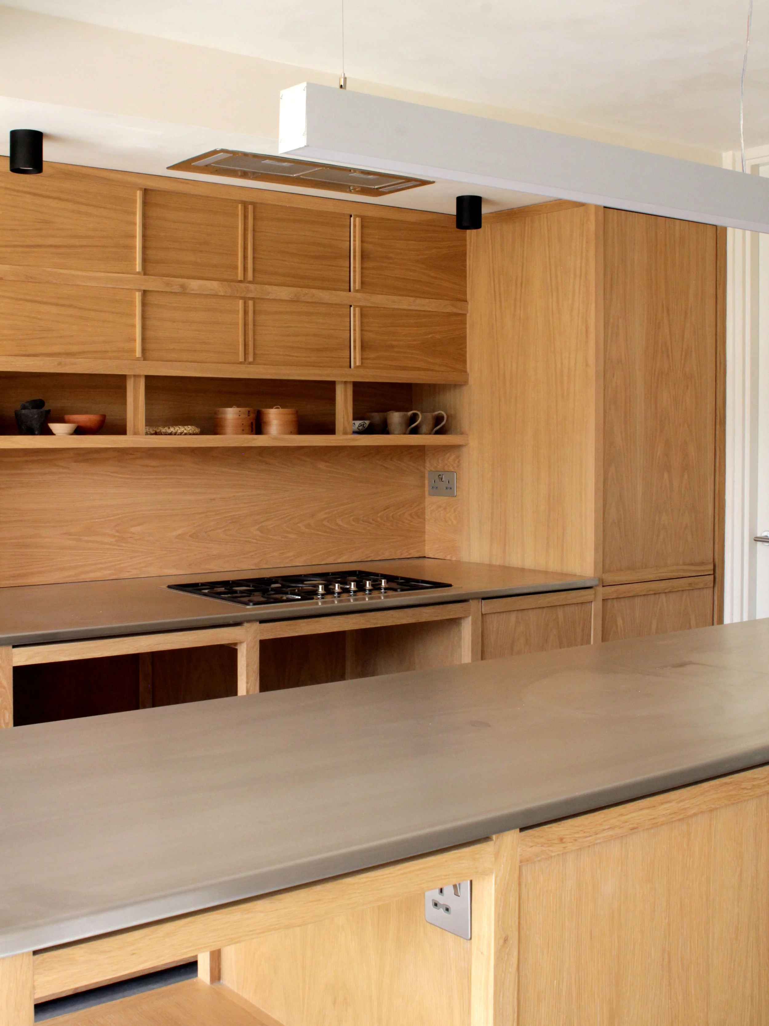 A kitchen with wooden cabinets, a countertop, and a stovetop, featuring open shelving with dishes and cups, and electrical outlets on the wall.