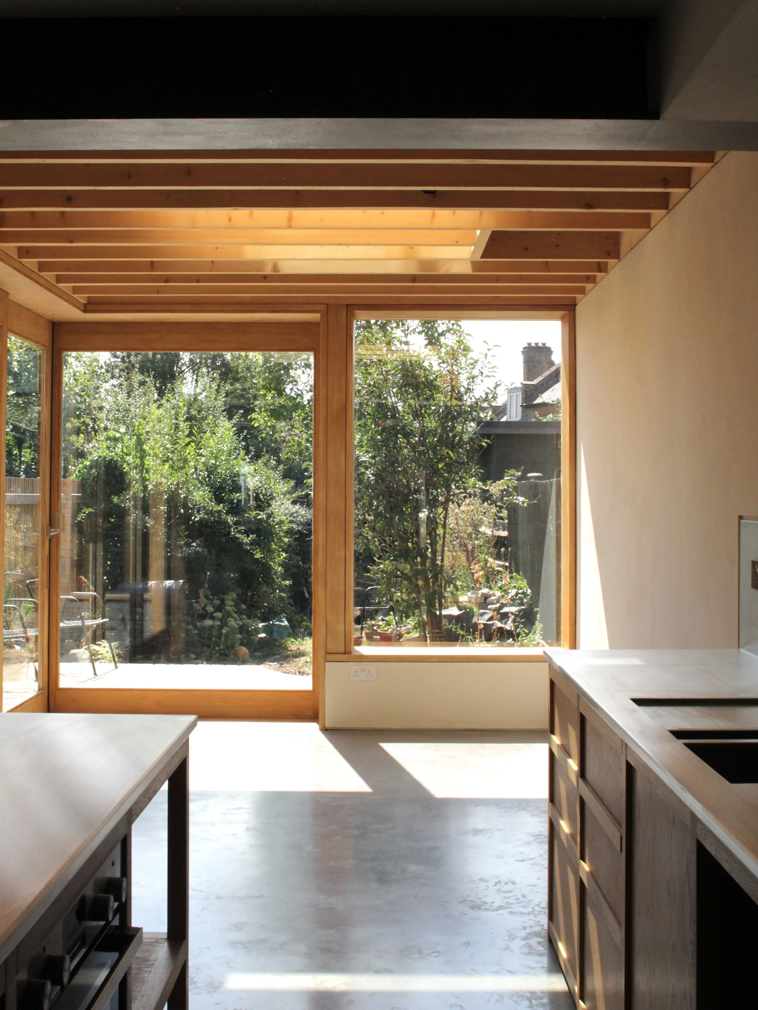 Kitchen with large glass sliding door and window overlooking a garden with trees and plants, wooden ceiling beams, light-colored walls and concrete floor, kitchen counter and cabinets on the right side.