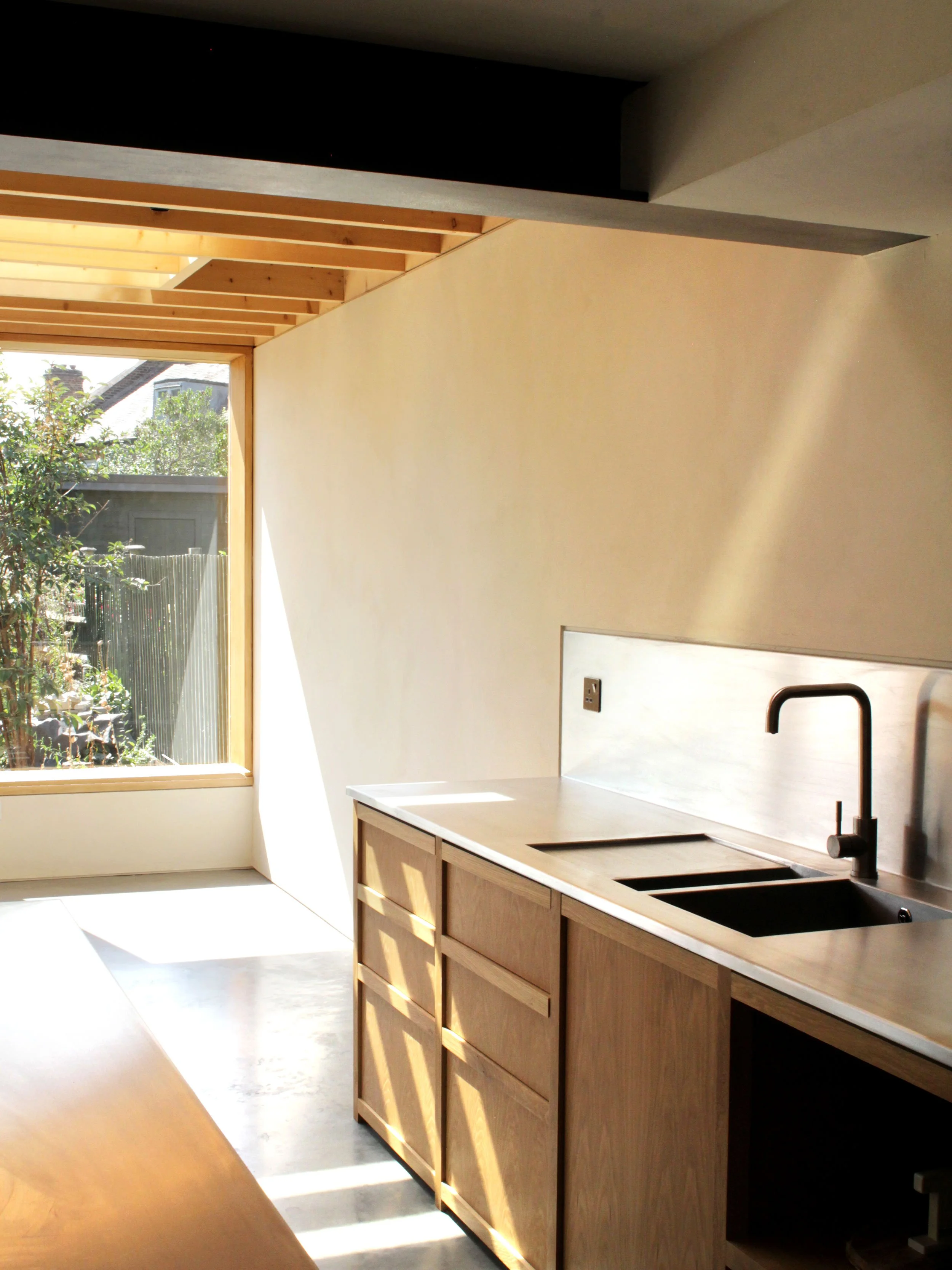 Interior view of a modern kitchen with wooden cabinets, a sink with a black faucet, and a large window allowing natural sunlight to illuminate the space.