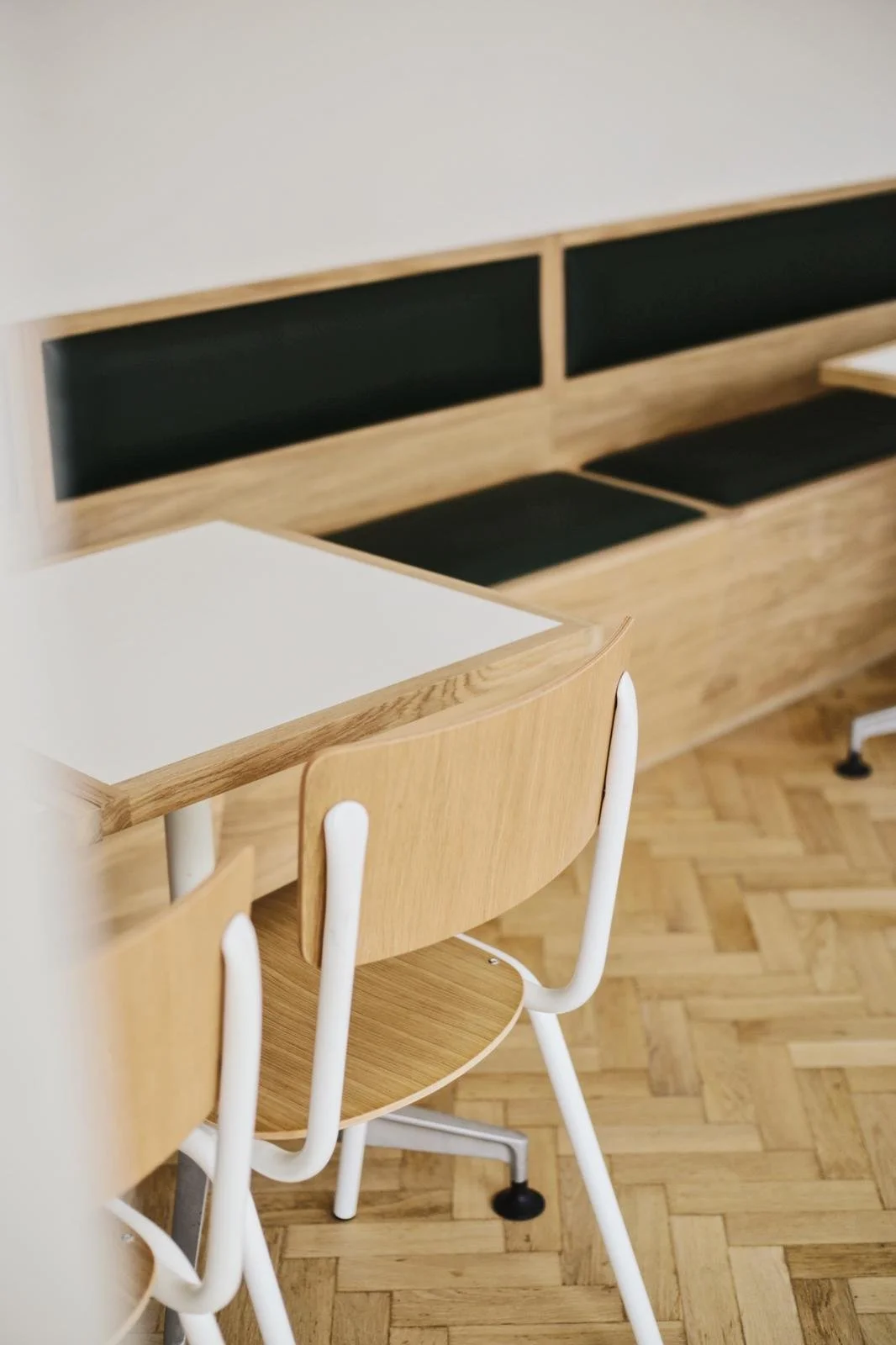 A wooden chair with white metal legs positioned next to a wooden table with a white top, in a room with a wooden floor, and a built-in wooden bench with black cushions along the wall.