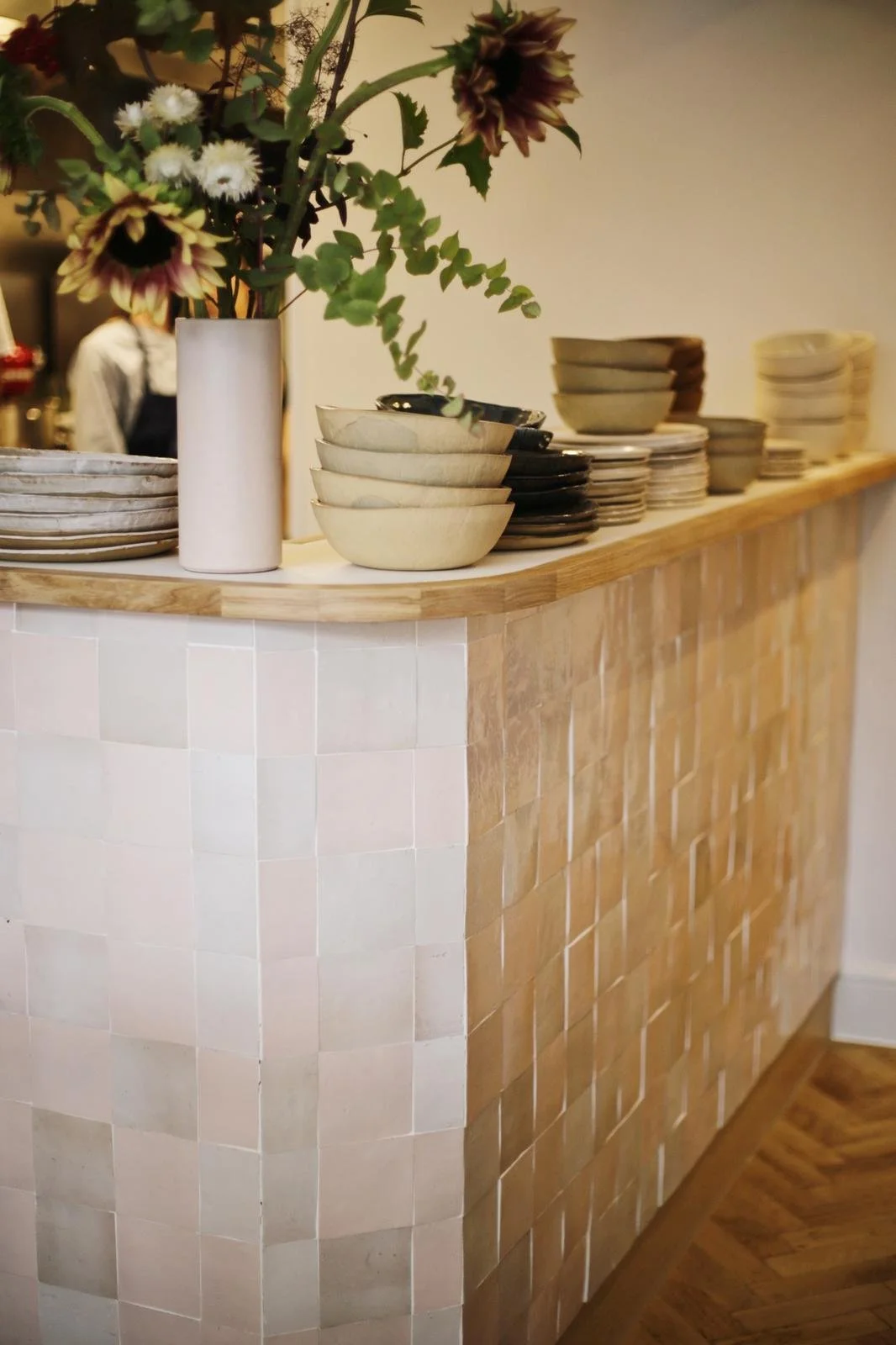 Stacked beige and black ceramic bowls and plates on a wooden counter, with a white vase of flowers and green foliage, in a restaurant or dining area.
