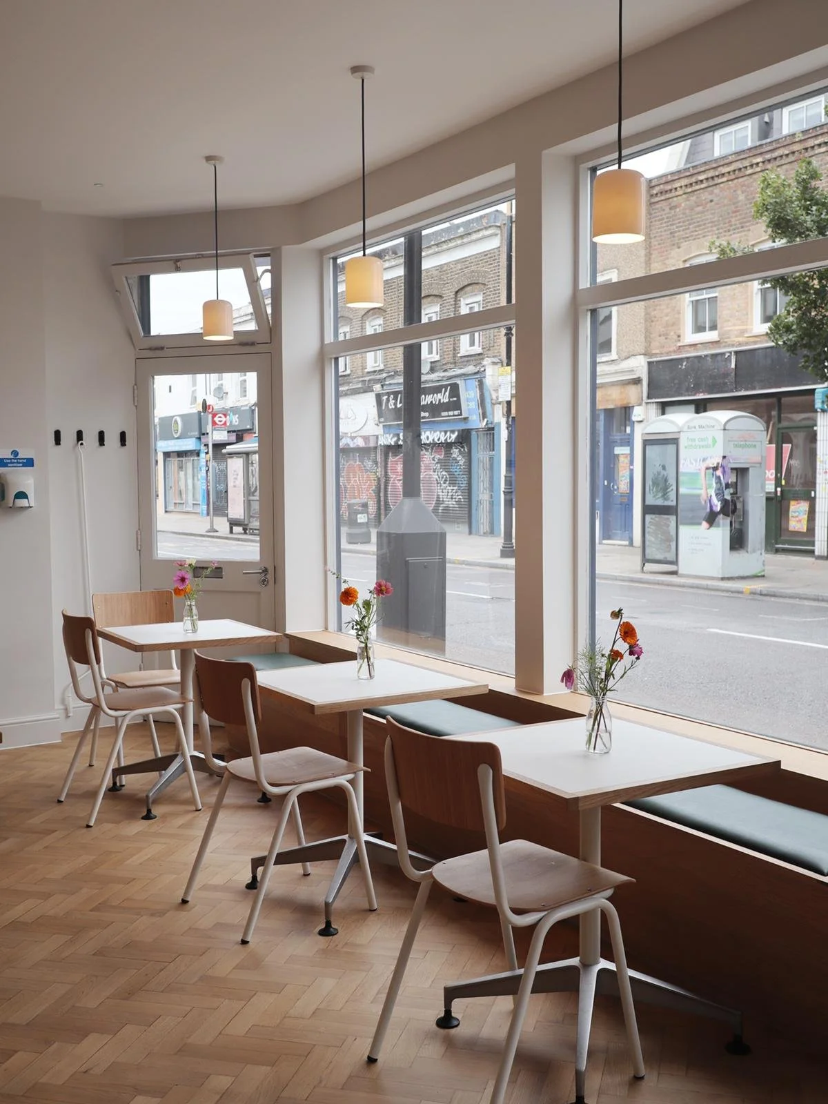 A cozy cafe with wooden tables, white chairs, and small vases of pink and orange flowers, illuminated by hanging pendant lights, with large windows showing an urban street scene outside.