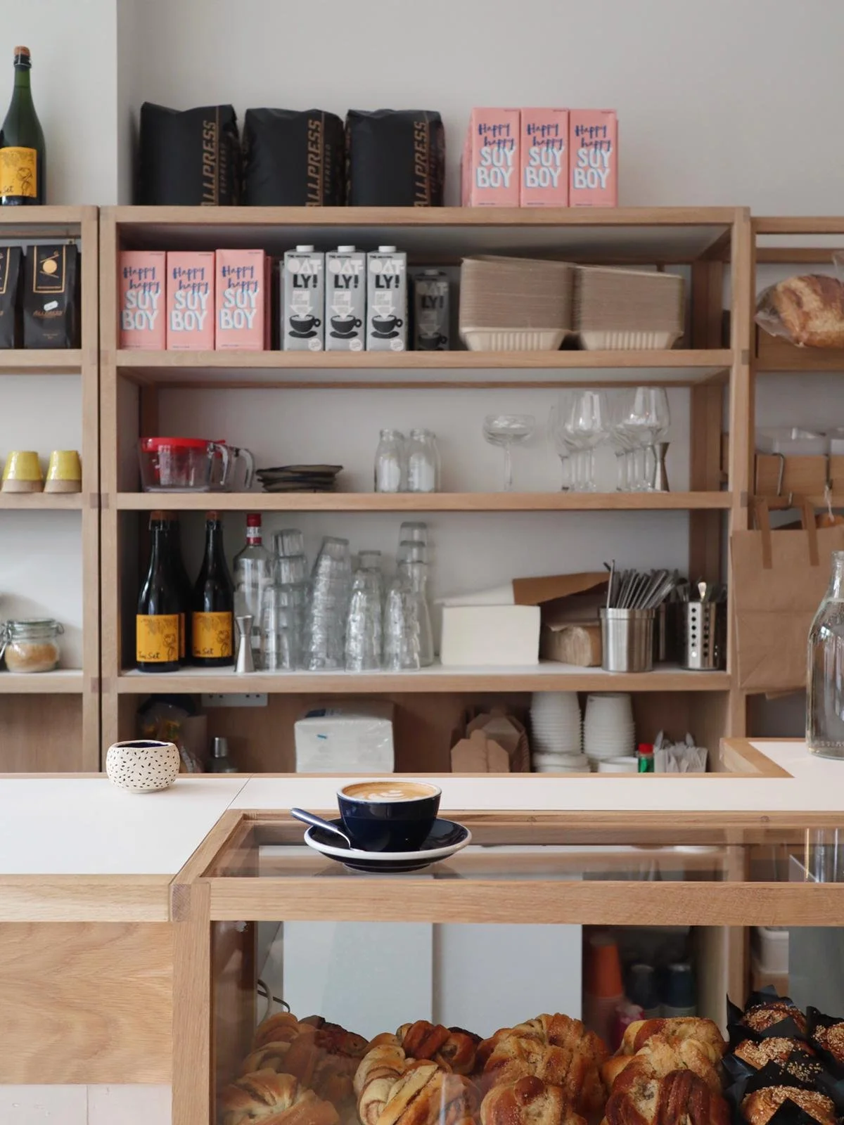 A coffee shop counter with a black coffee in a navy blue cup and saucer on a wooden stand, pastries behind a glass display case, and wooden shelves in the background holding glasses, drink cartons, bottles, and coffee supplies.