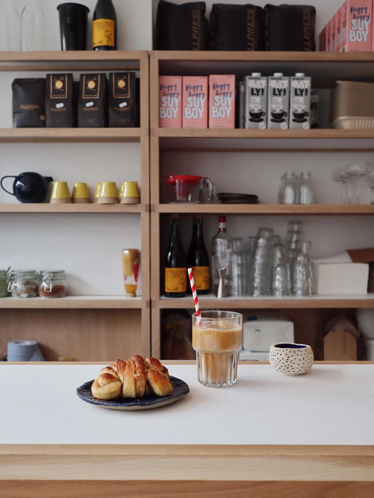 A cinnamon roll on a black plate, a glass of coffee with a red and white striped straw, and a small cup on a light-colored counter in front of wooden shelves with coffee cups, jars, bottles, and glasses.