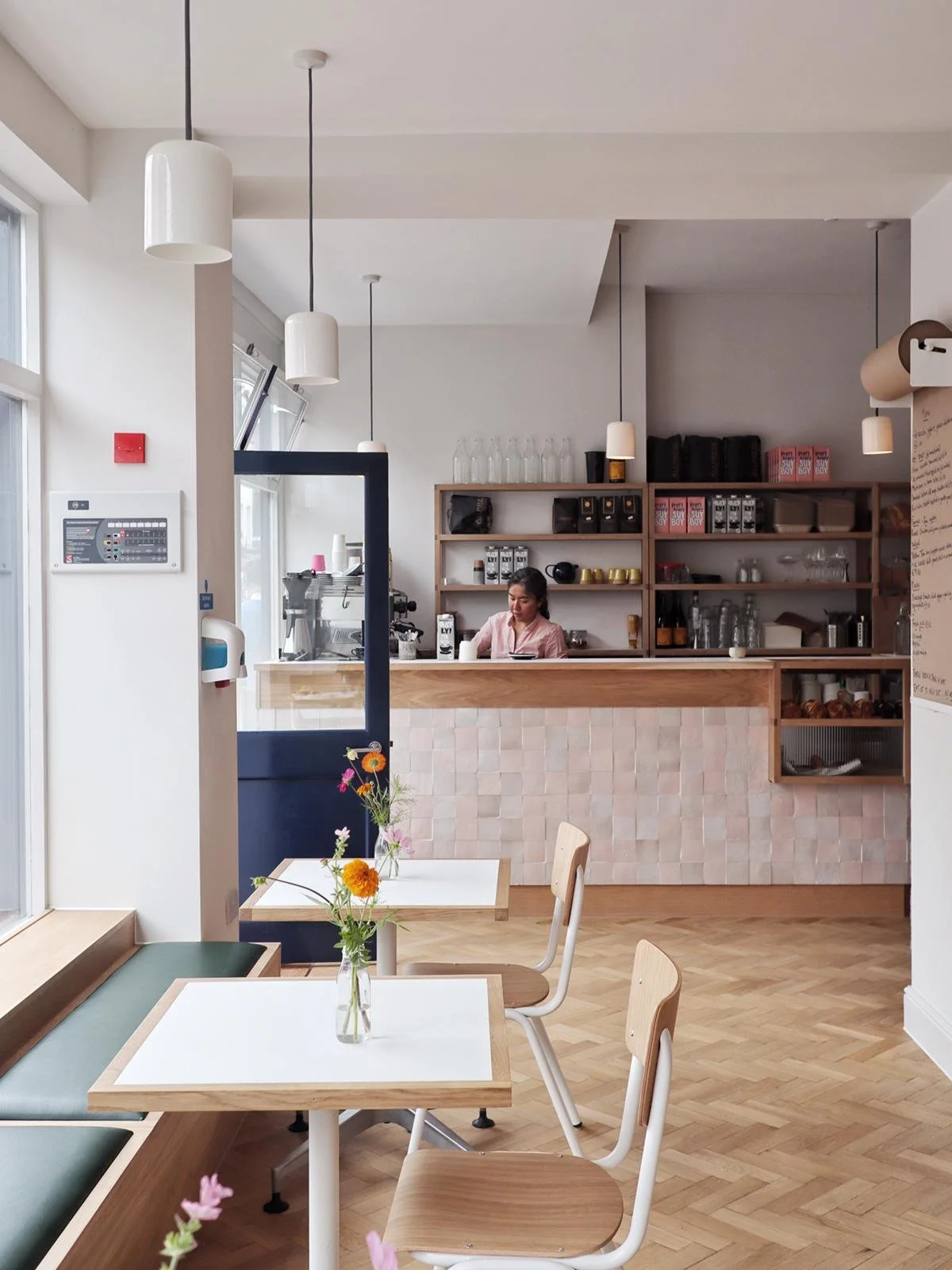 Interior of a modern cafe with white walls, wooden floors, and minimalistic decor. There are two white tables with wooden chairs, one with a small vase of colorful flowers. In the background, a barista works behind a pink tiled counter, with shelves 