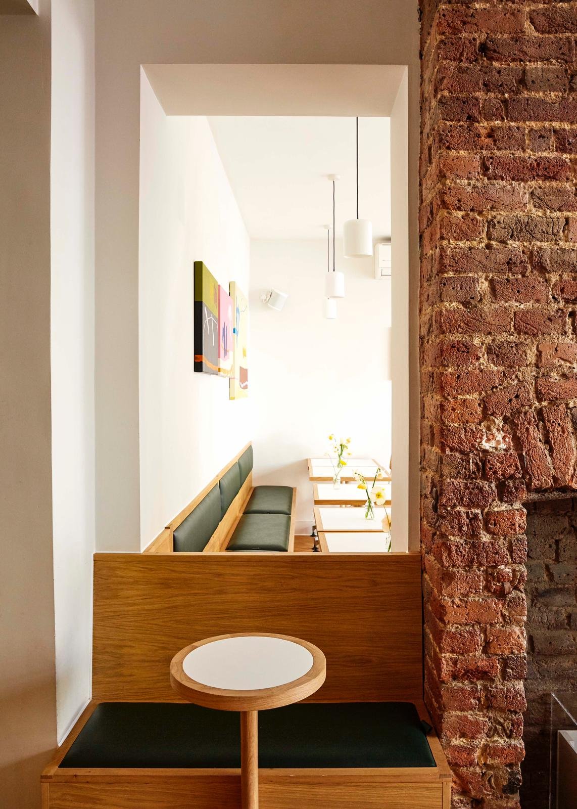 Interior view of a modern cafe or restaurant with a brick wall on the right, a white wall with colorful abstract art on the left, a green cushioned bench, and small tables with vases of yellow flowers, illuminated by hanging white pendant lights.