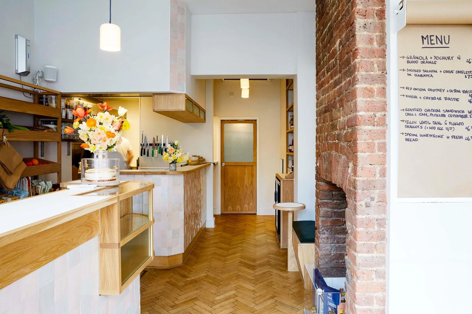Interior of a cafe with a wooden counter, flower vases, and a menu on the wall, brick and white walls, wooden flooring, and pendant lighting.