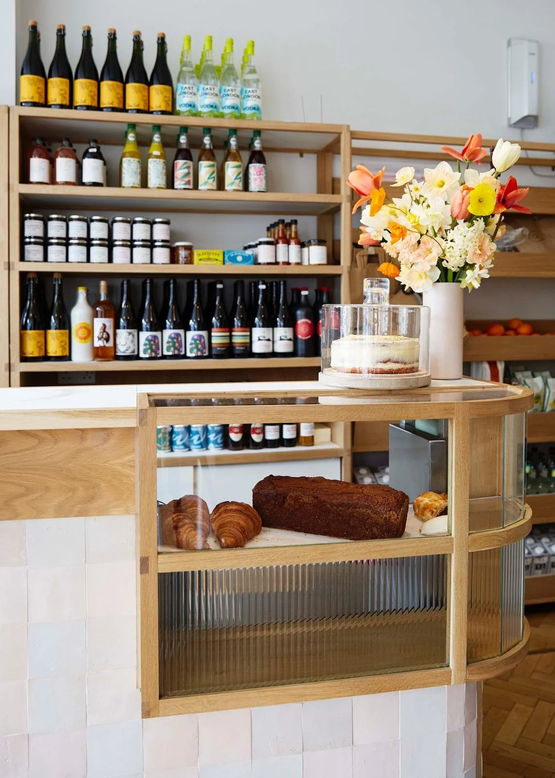 Display case with croissants, a loaf of bread, and a pastry and a cake on top of the counter, with a vase of flowers and shelves with bottled beverages and jars in the background.