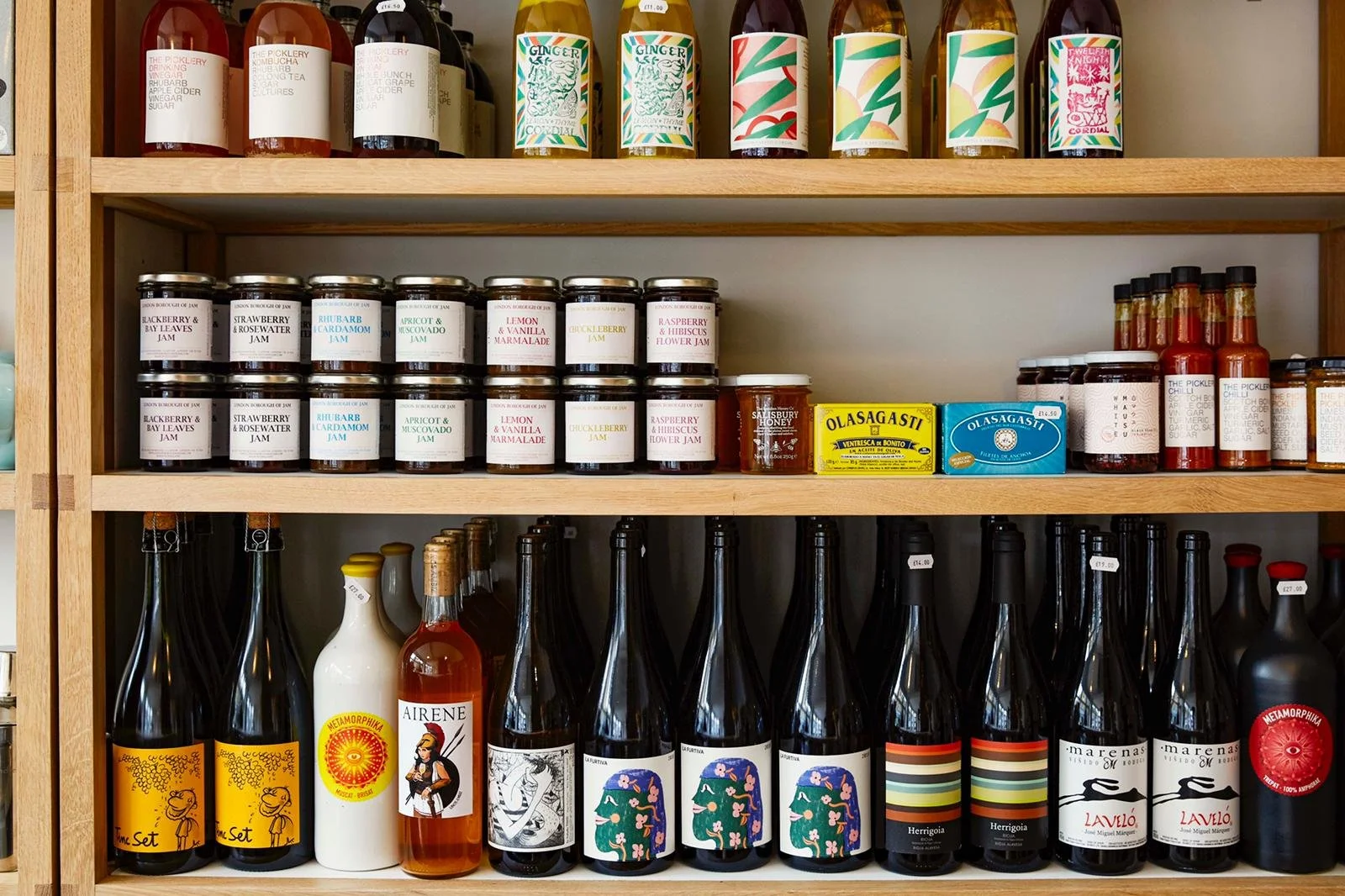 Shelves stocked with various bottles of beverages, jams, and condiments. Top shelf has colorful bottles of juice or soda. Middle shelf contains jars of fruit jams with labels such as 'Blackberry & Bay Leaves,' 'Strawberry & Rosewater,' and 'Lemon & V