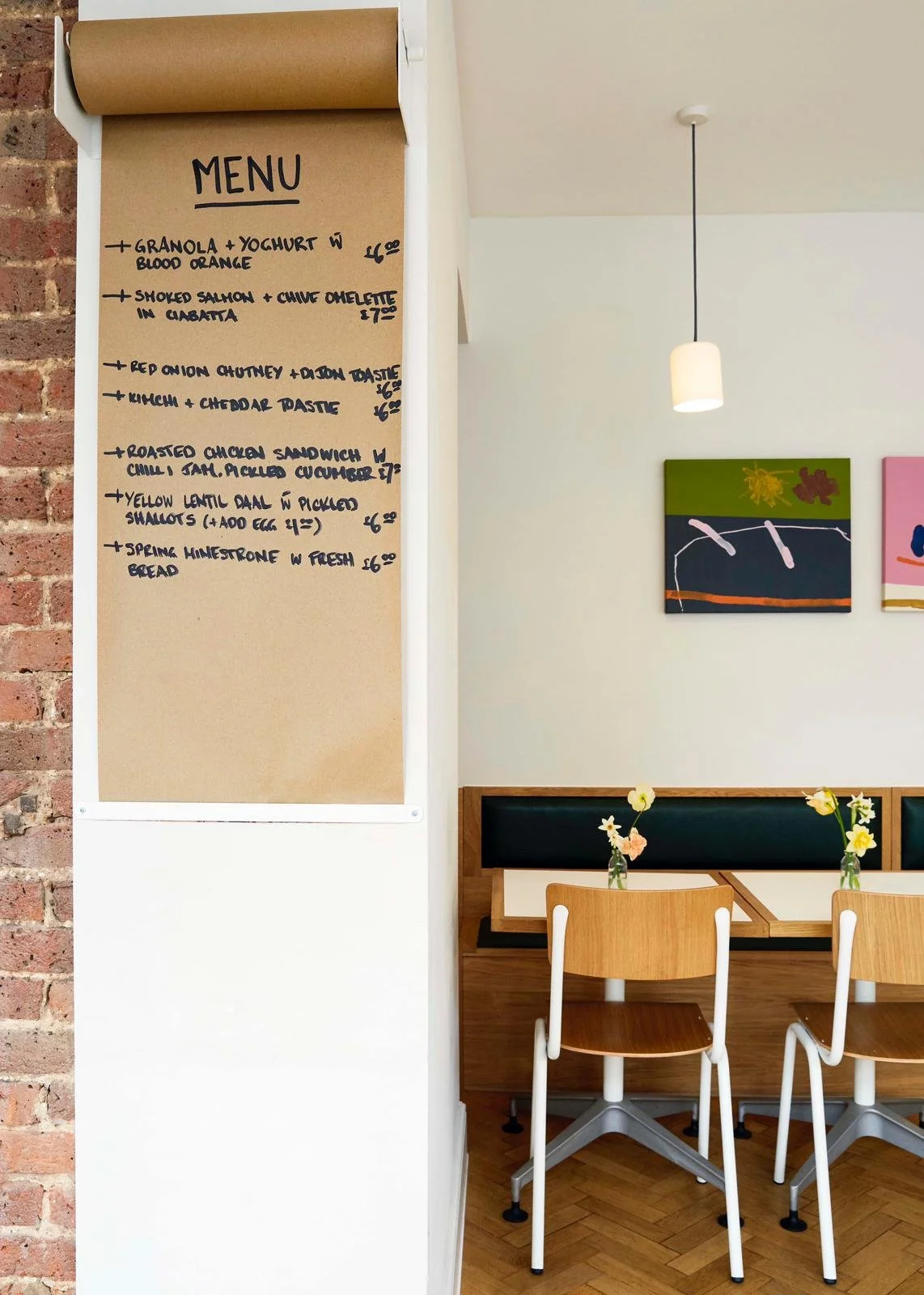 A restaurant menu written on a large brown paper scroll attached to a white wall, with six menu items listed. The restaurant interior features a lighting pendant, abstract paintings on the wall, and a seating area with wooden chairs and a table decor