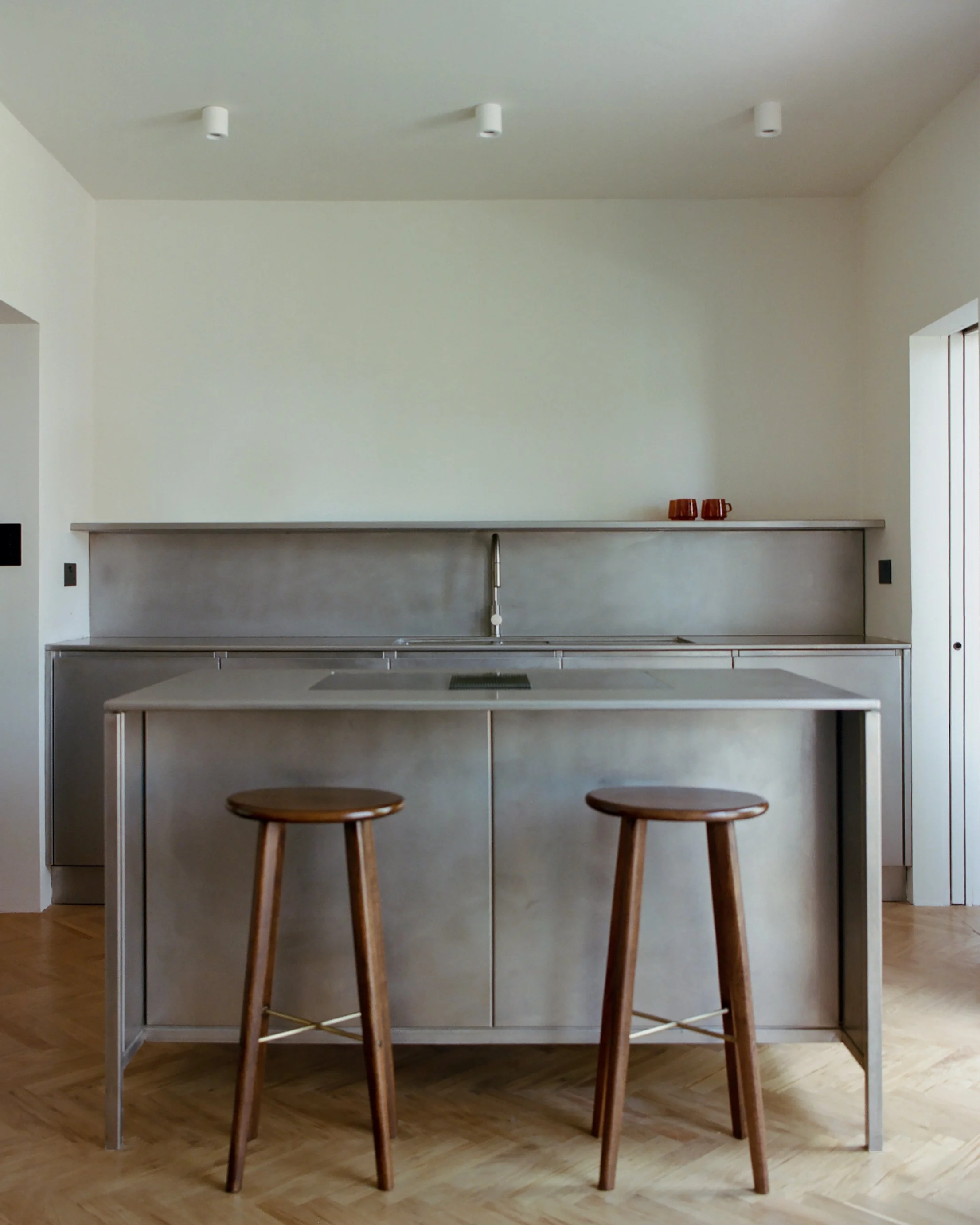 Minimalist kitchen with a stainless steel island and two wooden stools, white walls, and a small shelf with three red mugs.