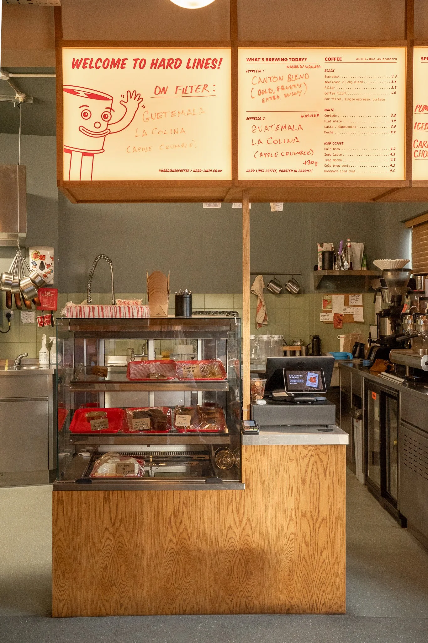 Coffee shop counter with a glass display case containing baked goods, a sink, and coffee equipment, with a menu board hanging above listing coffee options and brewing today.