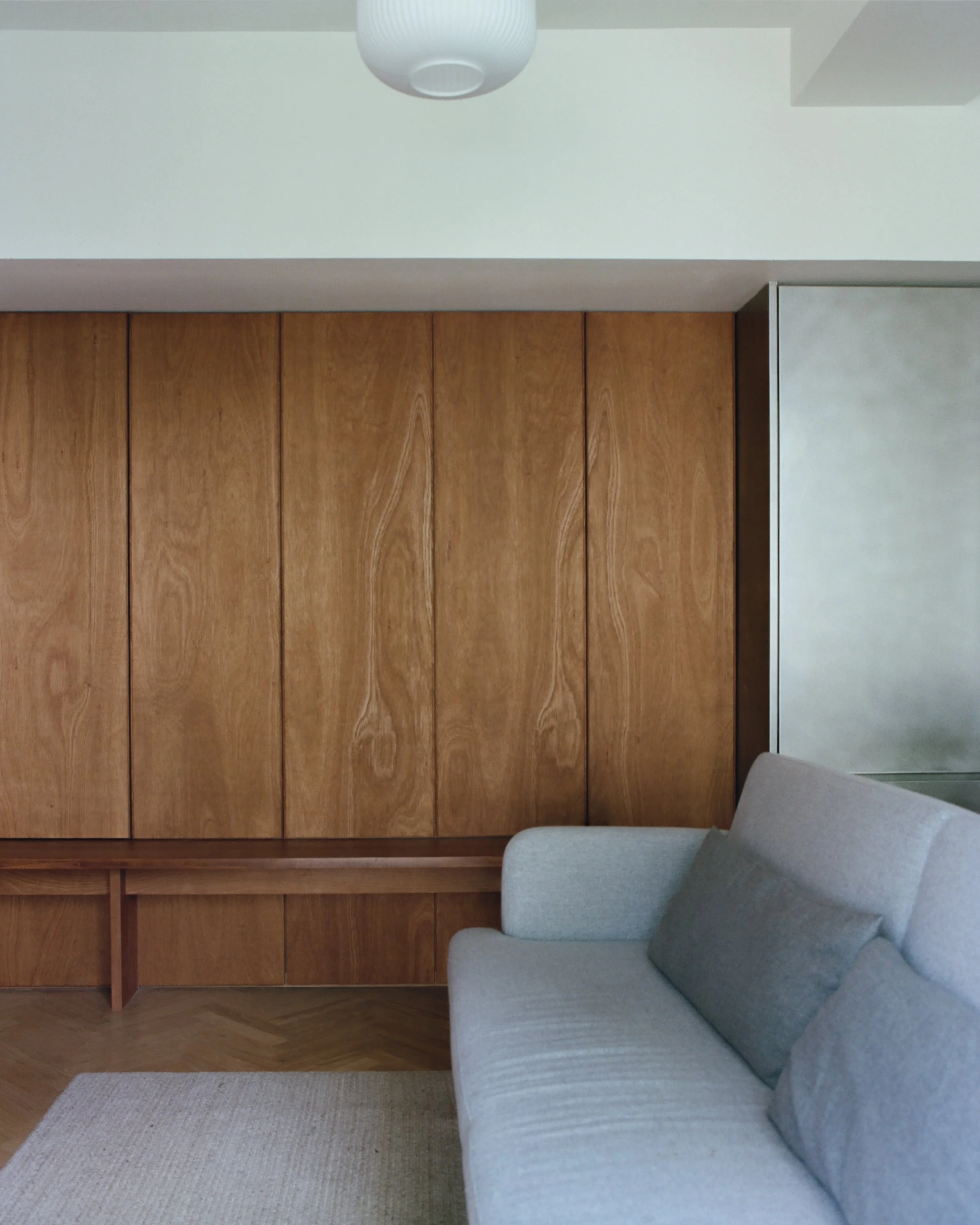 Living room with a wooden-paneled wall, a light gray sofa with darker gray pillows, a beige rug, a wood shelf, a chrome fridge, and a white ceiling light.