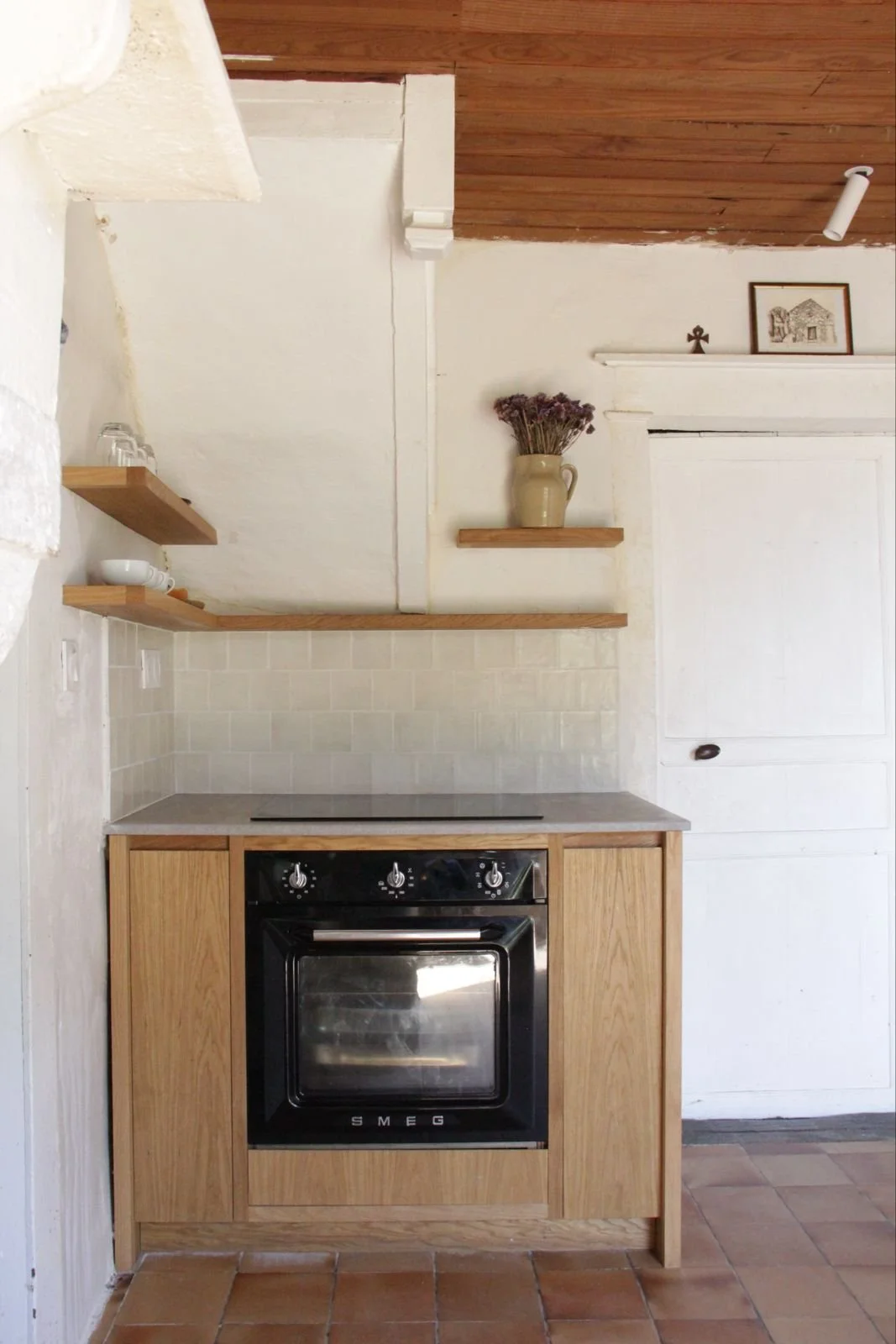 A kitchen with a vintage oven, wooden cabinets, and open wooden shelves with dishes and glasses, decorated with a vase of dried flowers and framed pictures on the wall.
