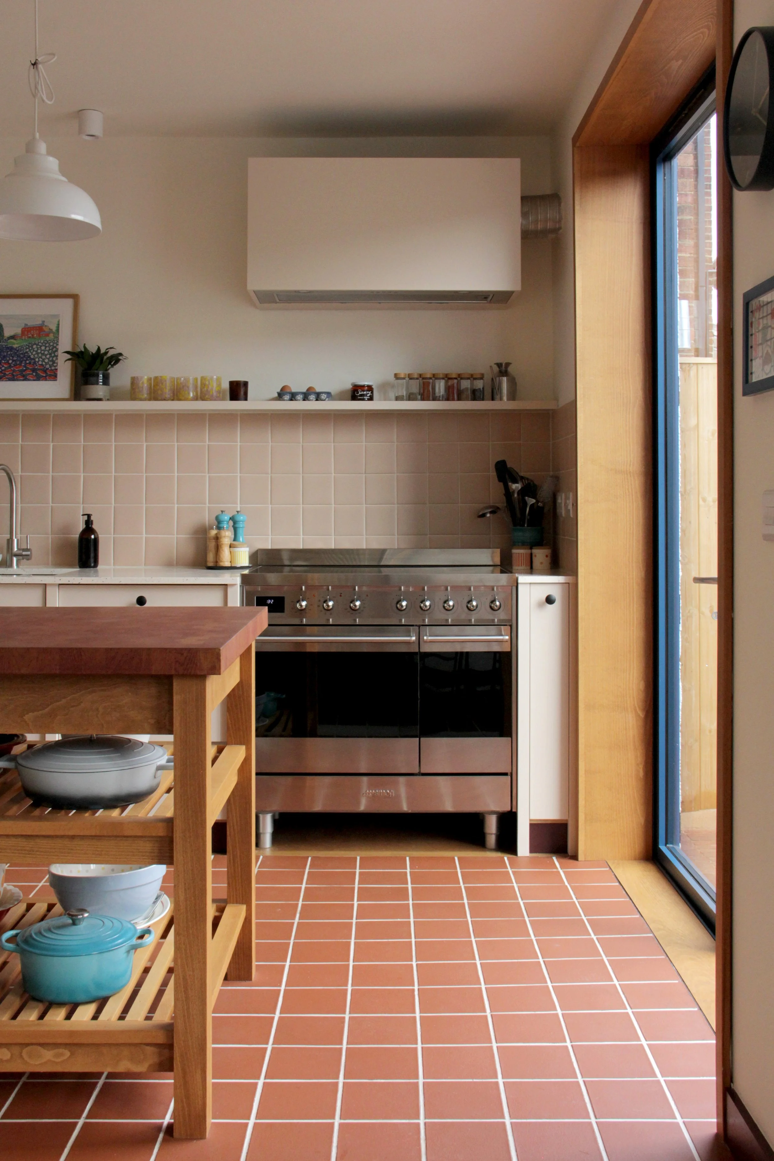 Kitchen with terracotta tile floor, wooden island, stainless steel stove, beige tiled backsplash, and a sliding glass door to the right.