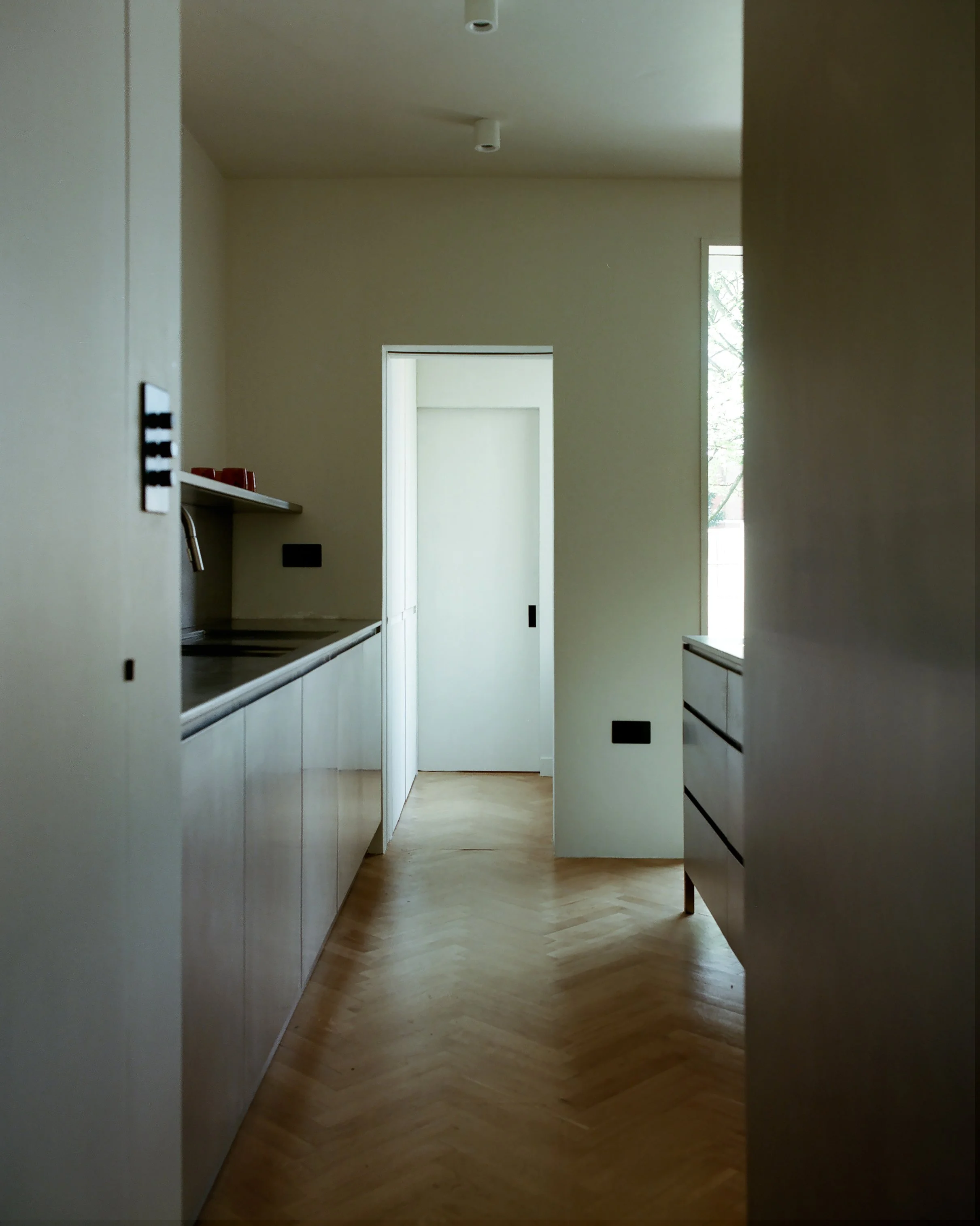 Empty modern kitchen with light wood cabinets, parquet flooring, and a doorway at the end of the room.