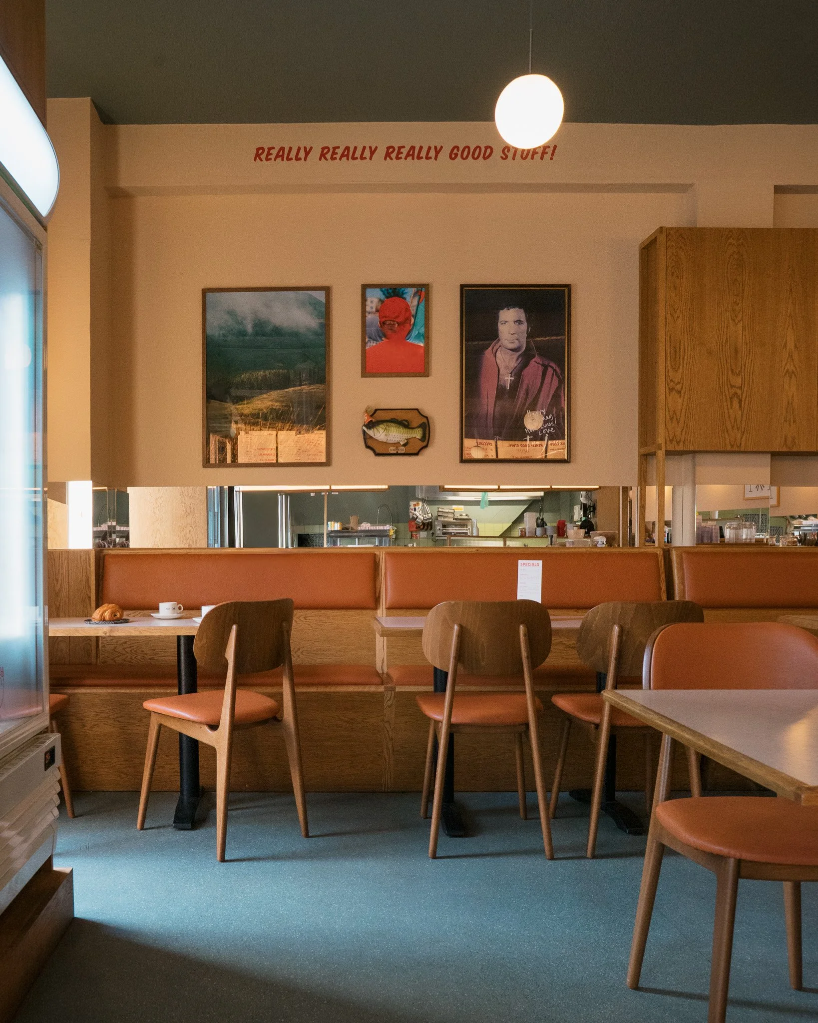 Empty dining area with wooden chairs and orange cushions, framed pictures on the wall, and a round light fixture hanging from the ceiling.