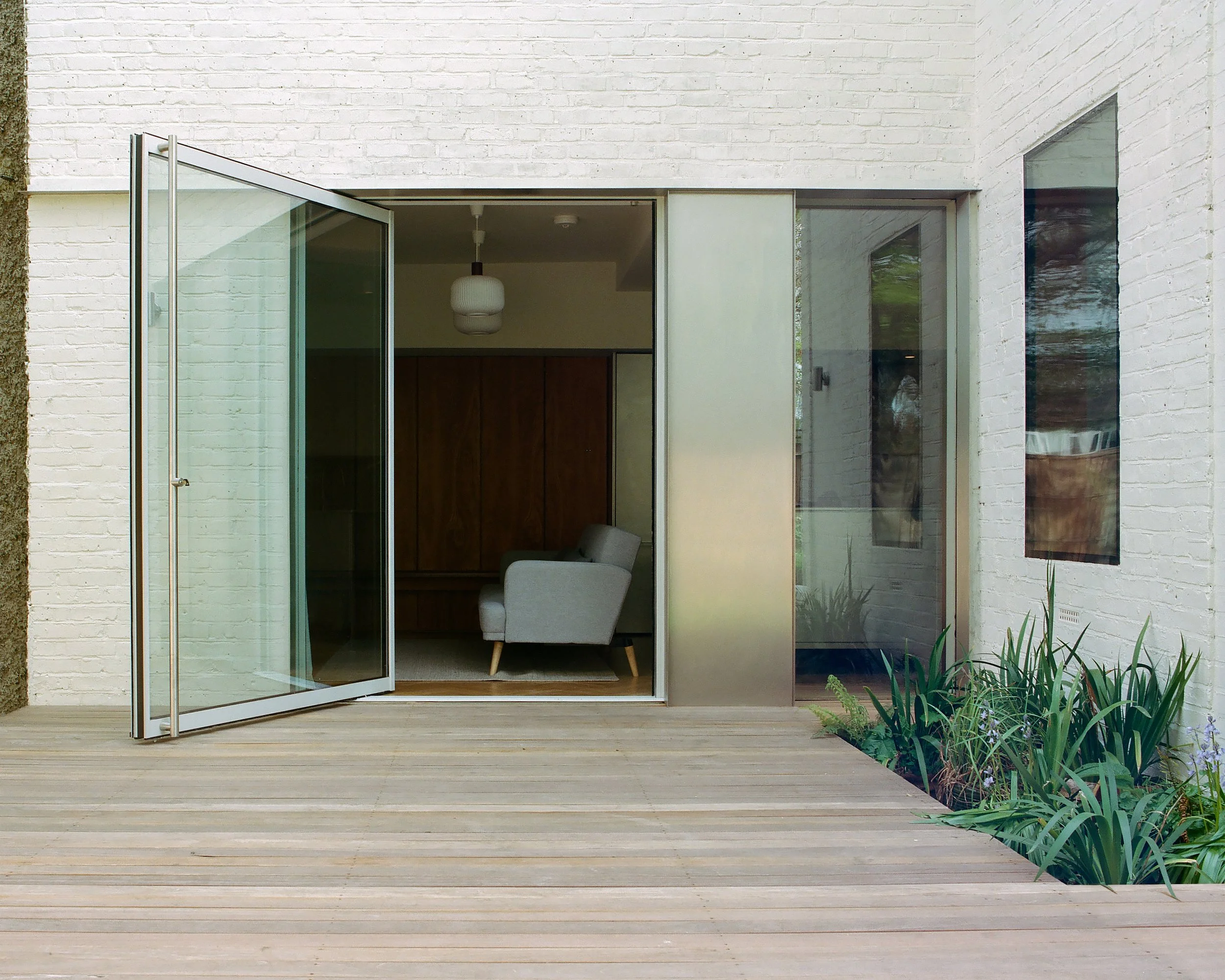 Modern white brick house with a wooden deck and sliding glass door leading to a living room with a chair and wooden cabinet.