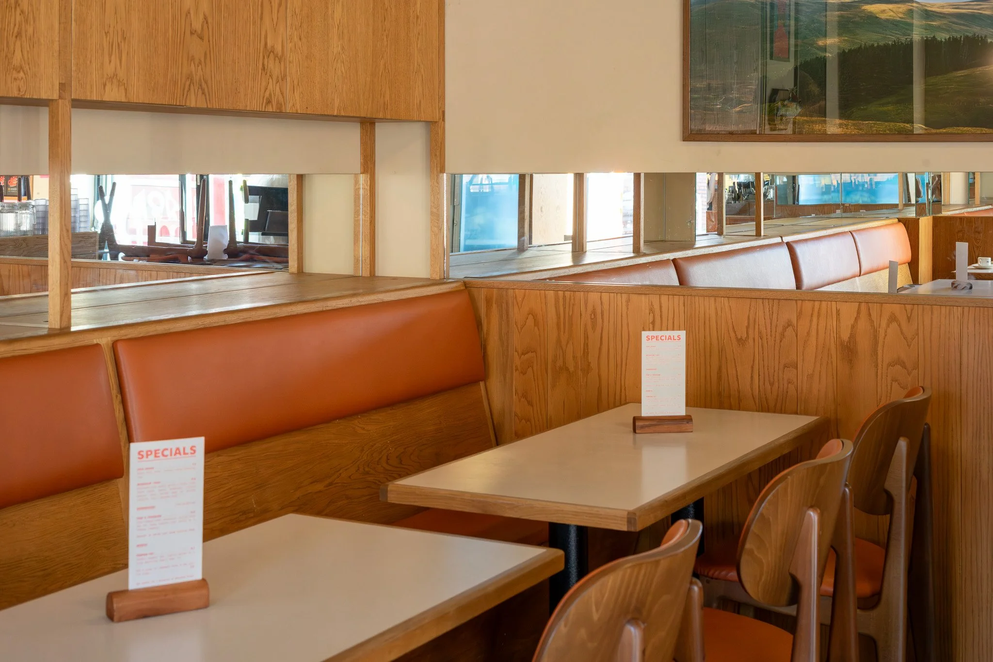 Empty restaurant booth with wooden walls, tables, and chairs, with menus on the tables and large windows showing a view outside.