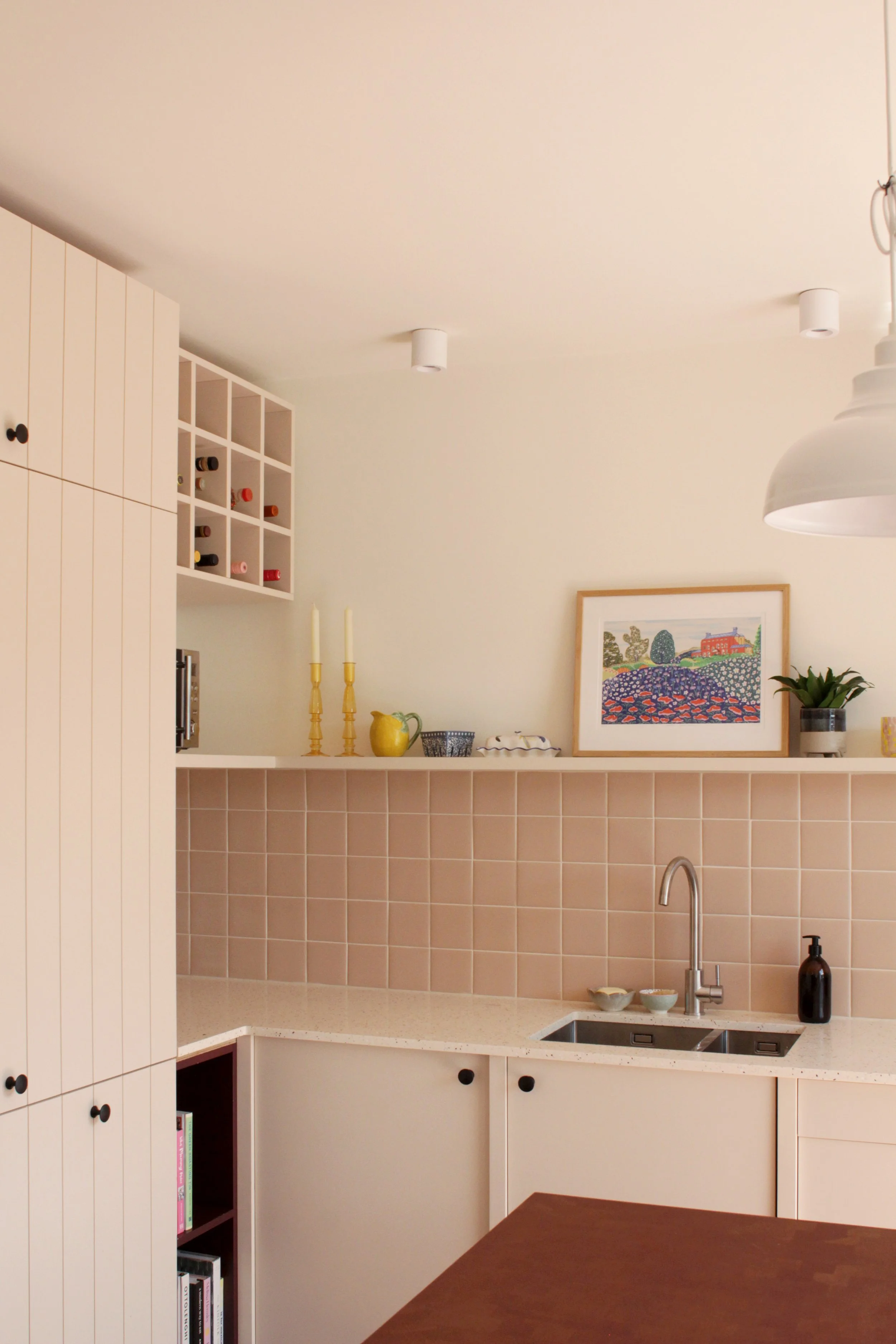 A modern kitchen with white cabinets, a tiled backsplash, and a white countertop. There are candles, a framed artwork, plants, and decorative items on the shelf above the sink.