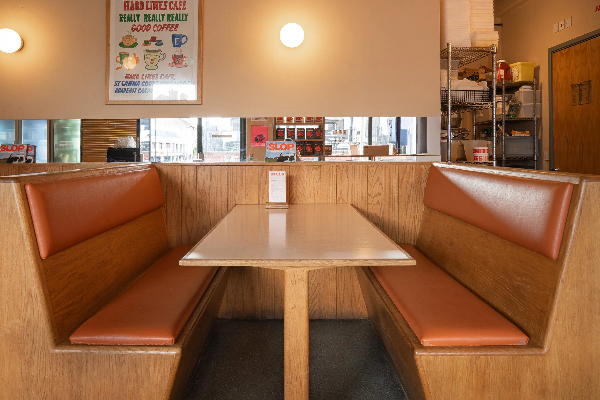 Empty booth seat at a wooden diner table with a menu stand, inside a cafe with wood-paneled walls, a large signboard, and a storage shelf in the background.