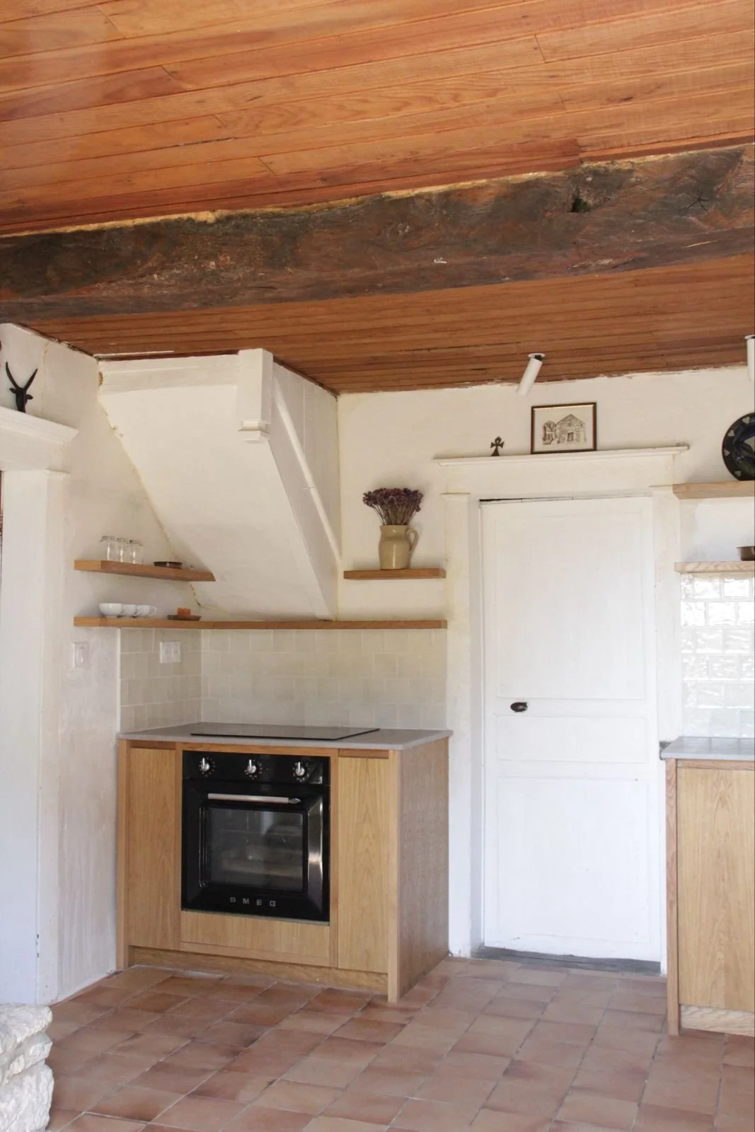 A rustic kitchen with a wooden ceiling and a small stove, white walls, open wooden shelves, and decorative items.