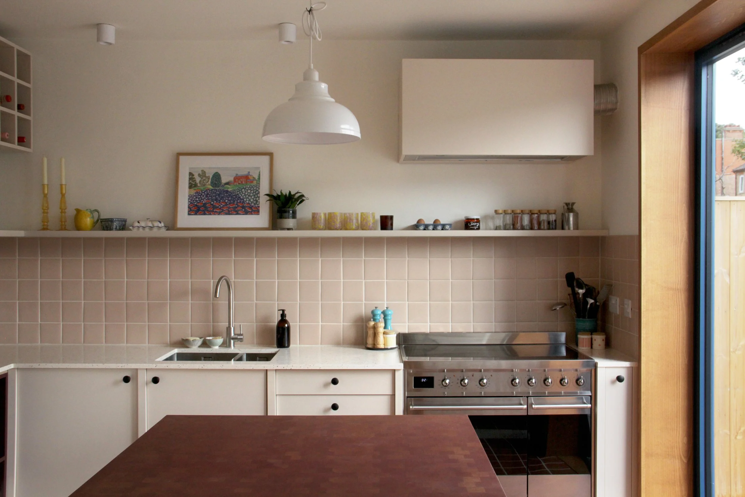 A modern kitchen with white cabinetry, beige tiled backsplash, stainless steel oven, a white countertop with a sink, and a window on the right side letting in natural light. The shelves above hold various small items and a framed colorful picture.