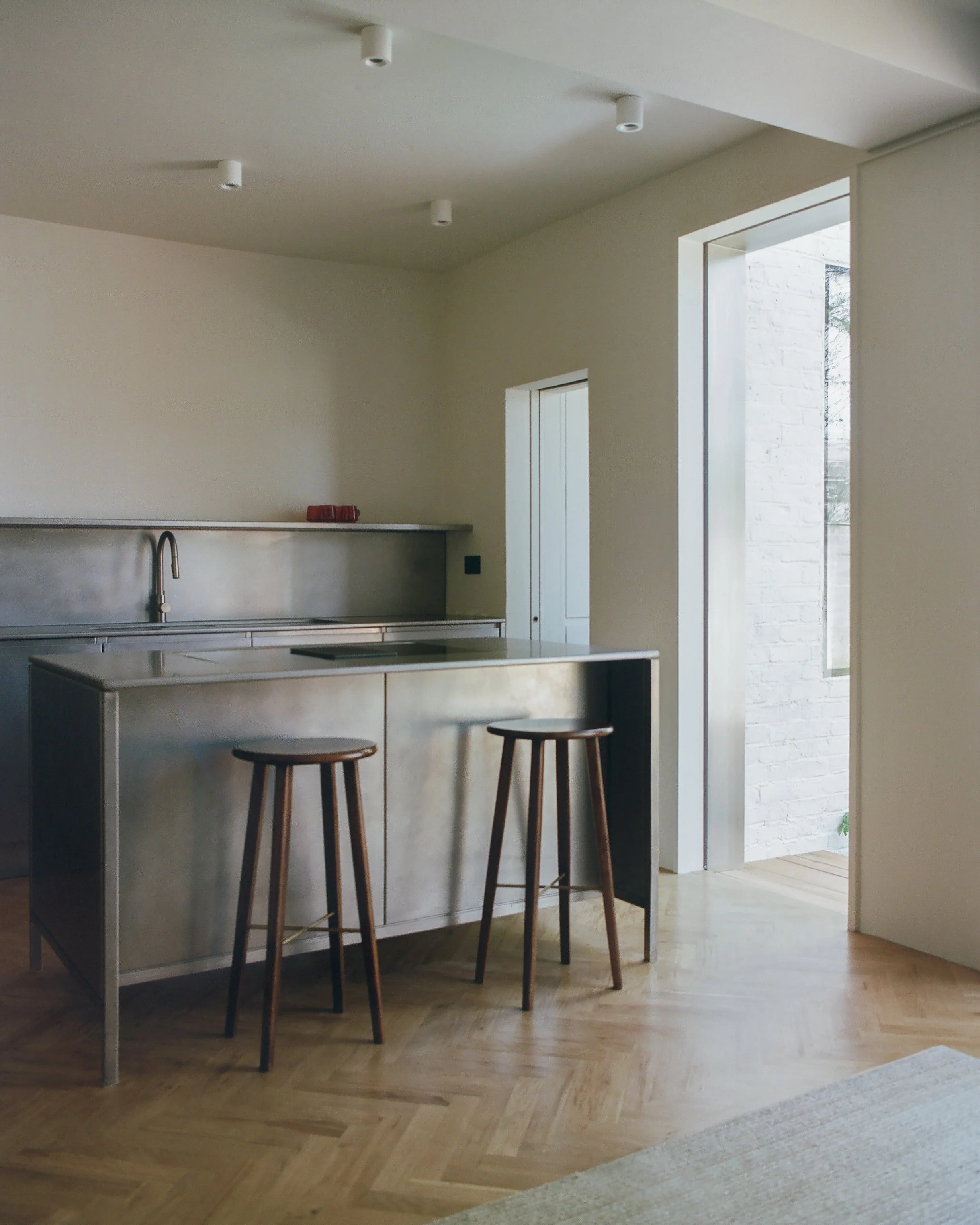 A modern kitchen with a stainless steel island counter and two wooden bar stools, with white walls and large windows letting in natural light.