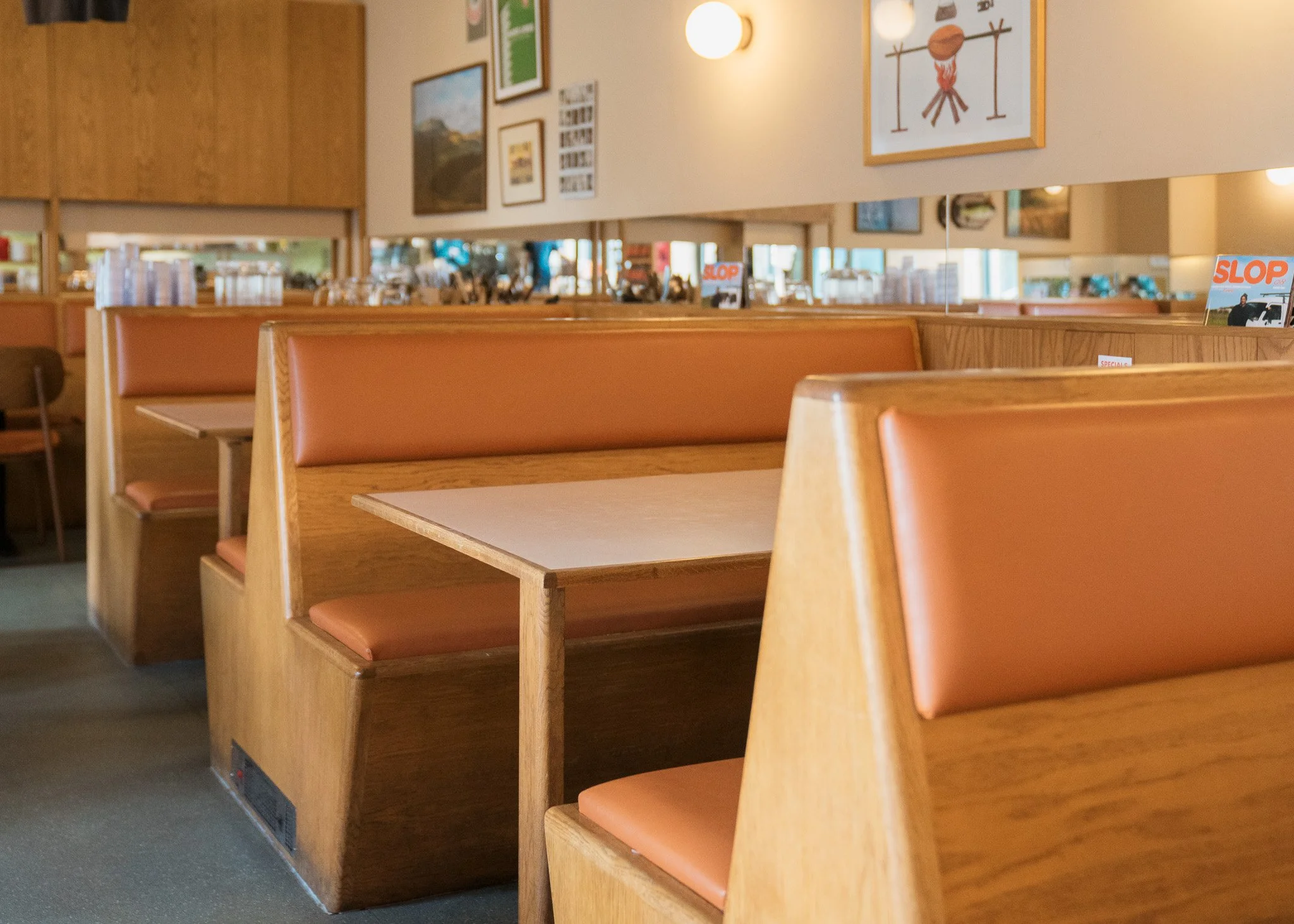 Empty booths with orange cushions and tables in a diner or restaurant setting, with wall art and mirrors in the background.
