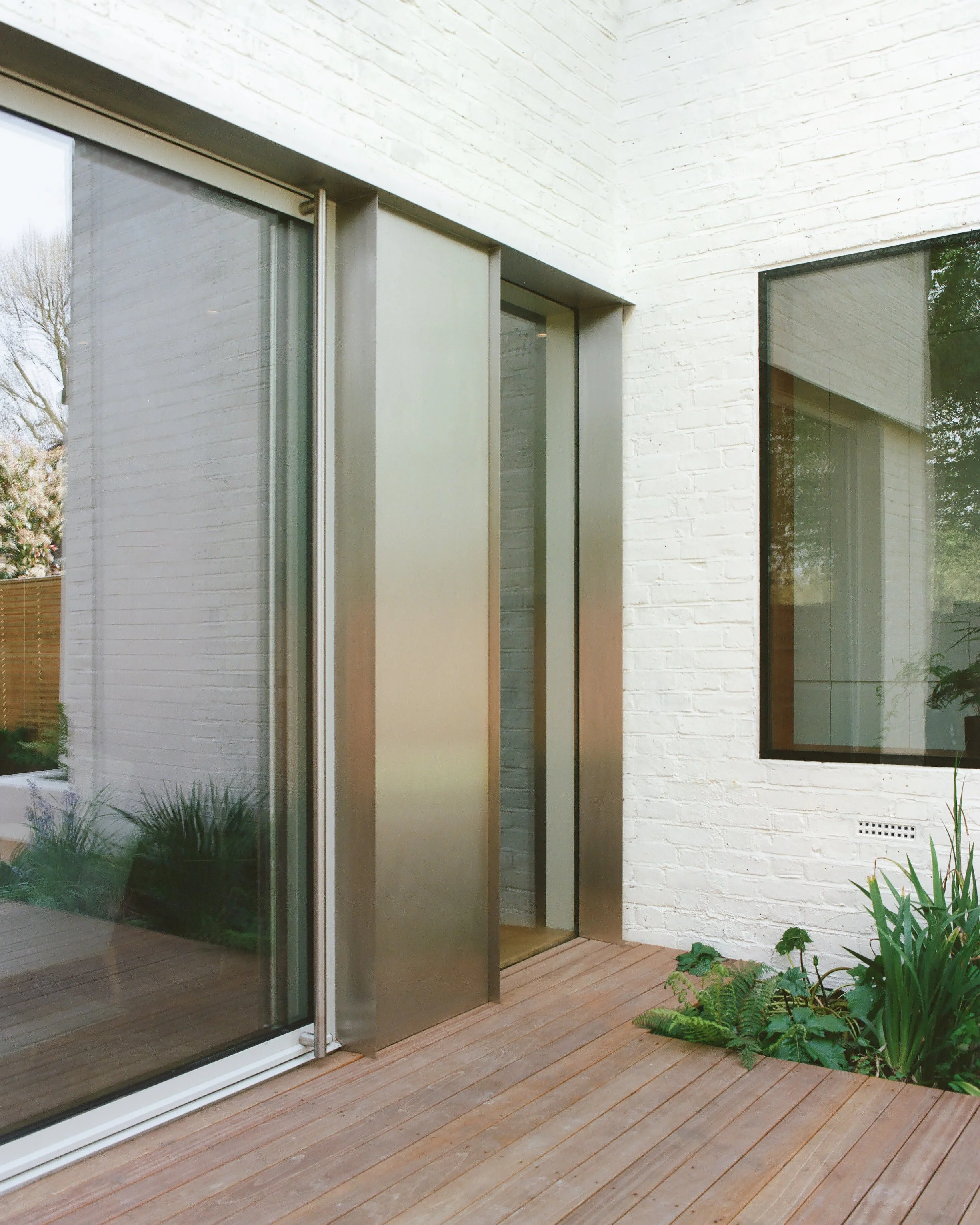Modern house exterior with large glass sliding door, white brick walls, and a wooden deck with some green plants.