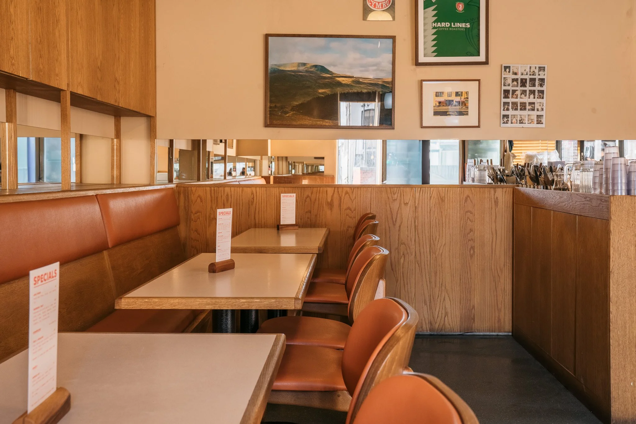 Interior of a cozy café with wooden paneling, brown leather chairs, framed landscape photographs and artwork on the walls, and a row of tables with small menus on stands.