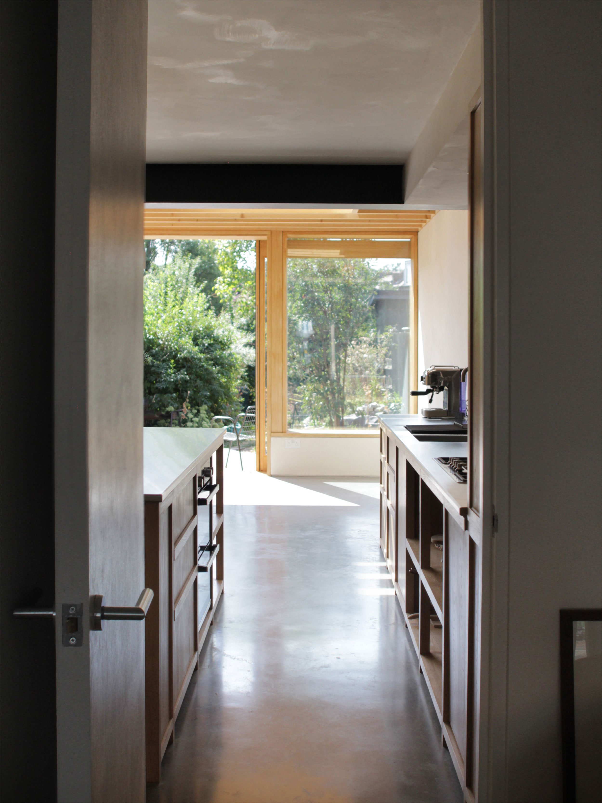View through doorway into a bright, modern kitchen with large window showing trees outside, wooden cabinets, and a concrete floor.