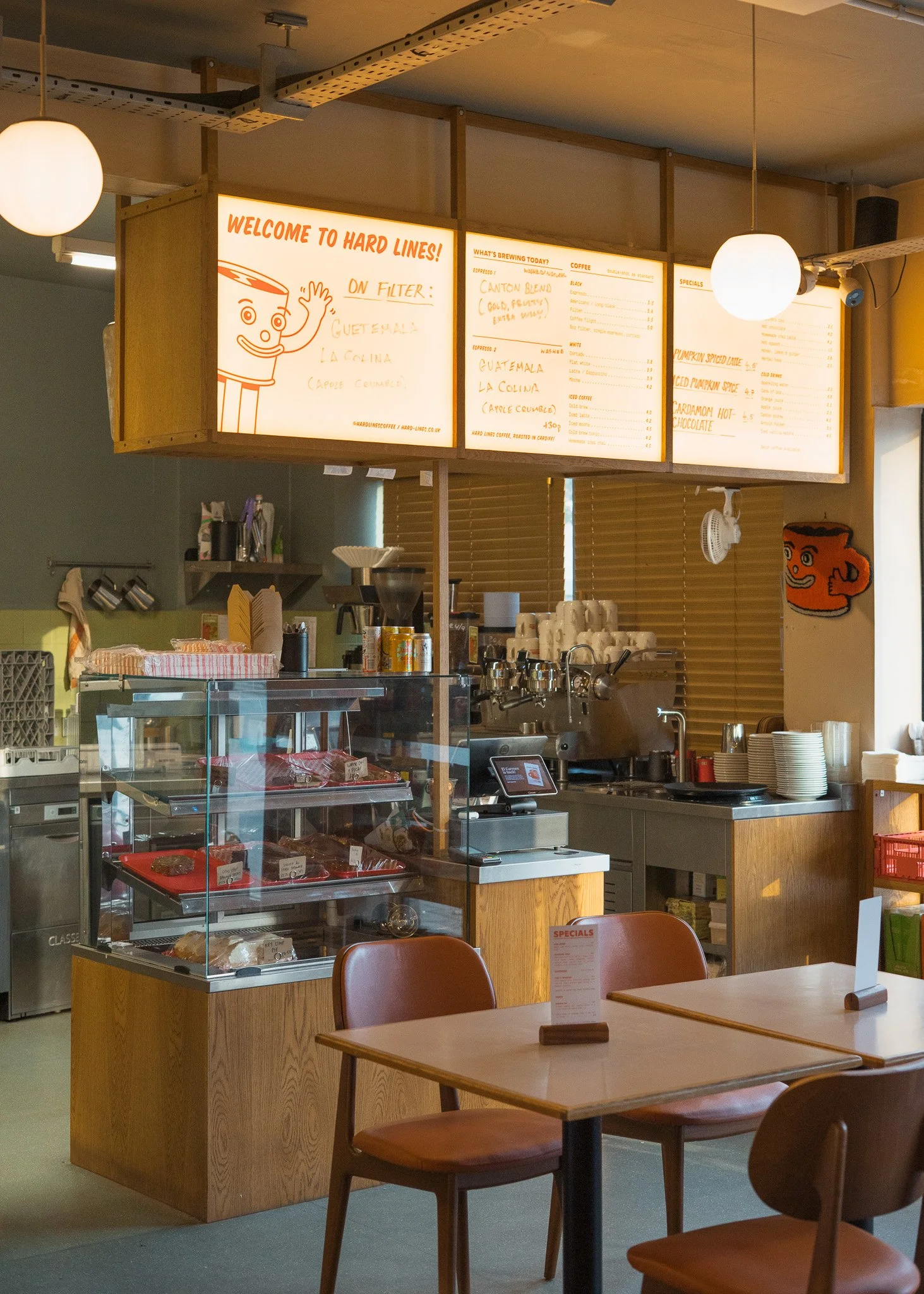 Inside a coffee shop with a counter displaying baked goods, a menu board above with coffee options and specials, and seating area with wooden tables and chairs.
