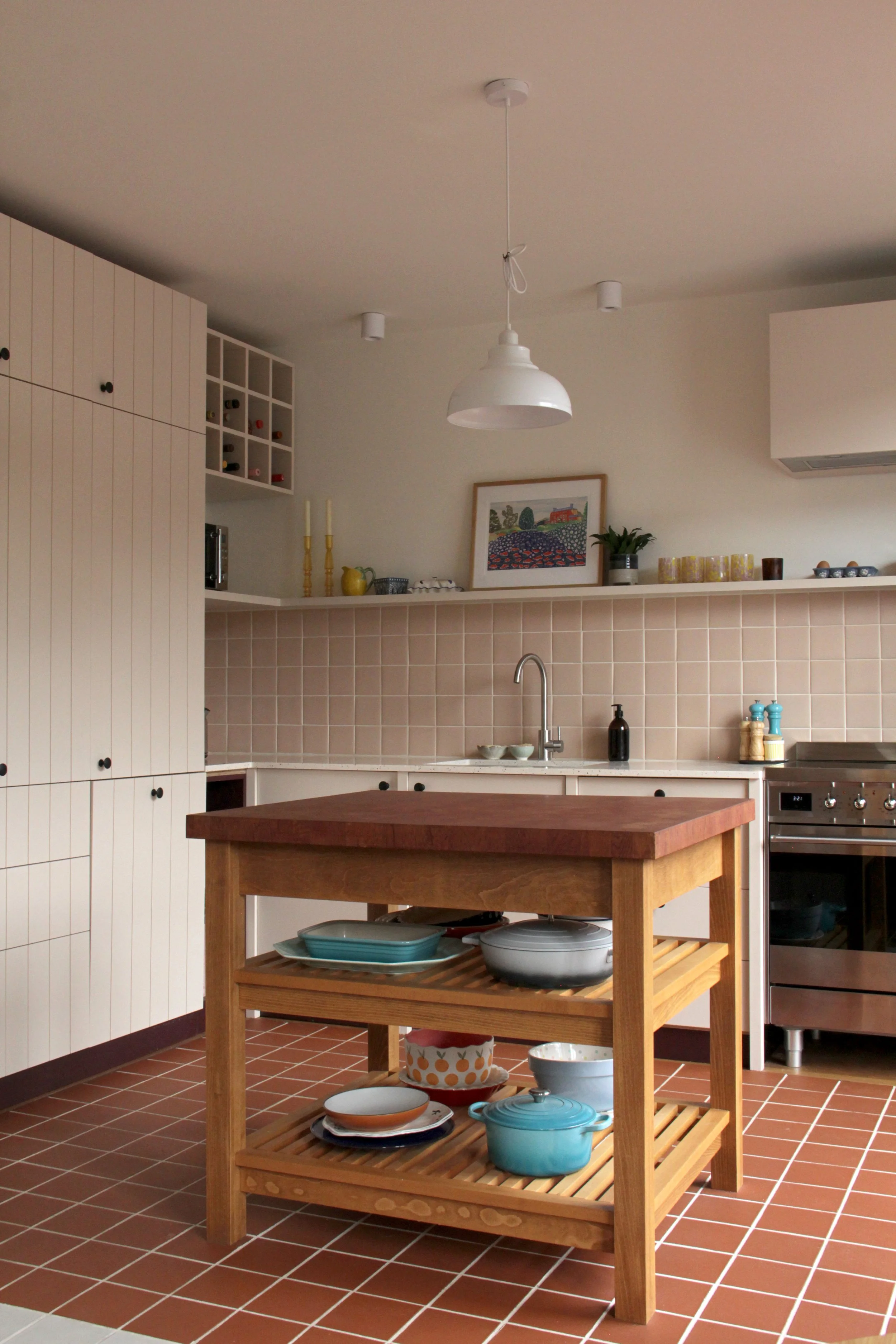 A modern kitchen with white cabinets, a wooden island, pink tiled backsplash, and a stainless steel stove. Decor includes framed artwork, potted plants, and yellow candles on a shelf.
