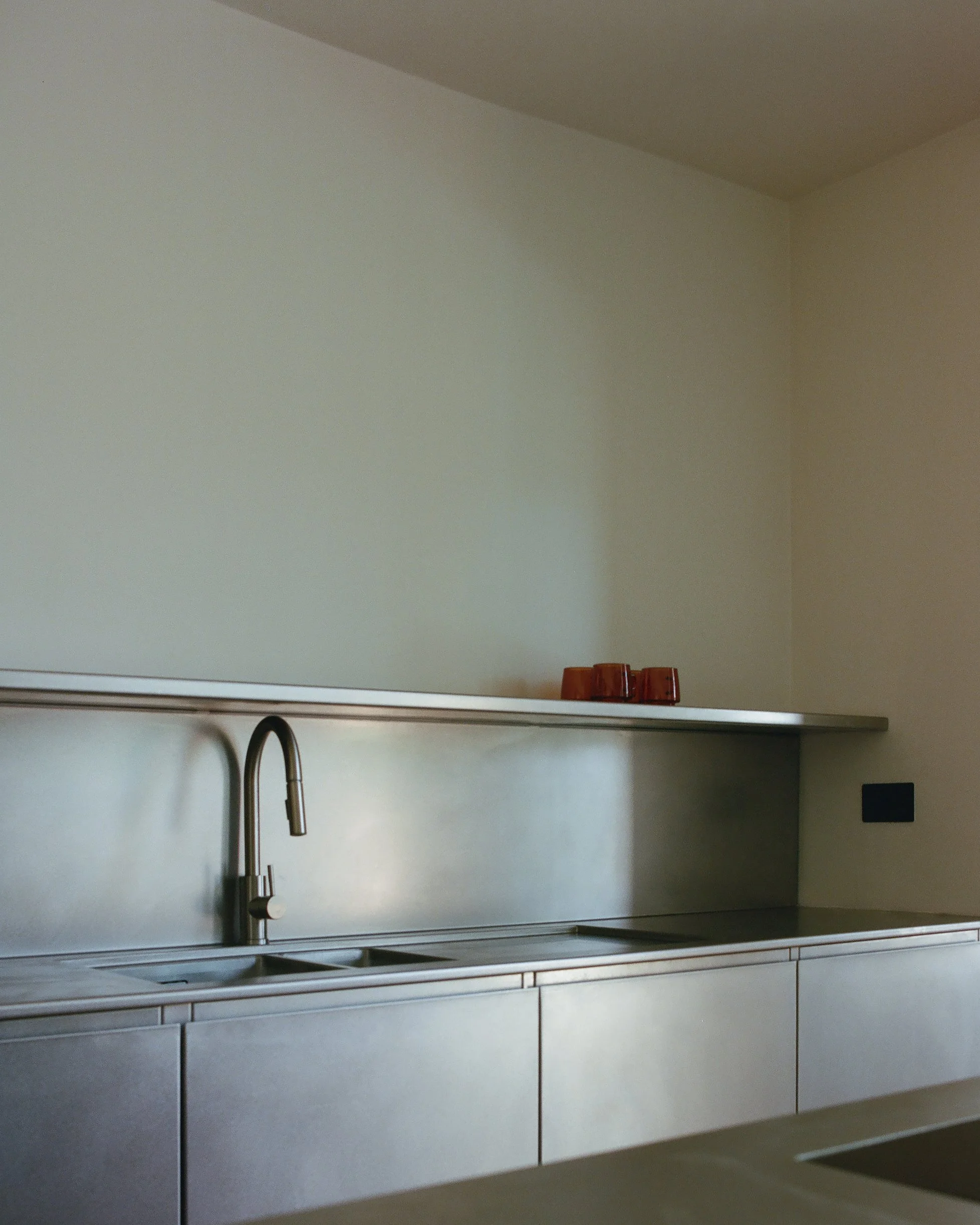 Minimalist kitchen with stainless steel sink and countertop, brown cups on shelf, and a dark electrical outlet on the wall.