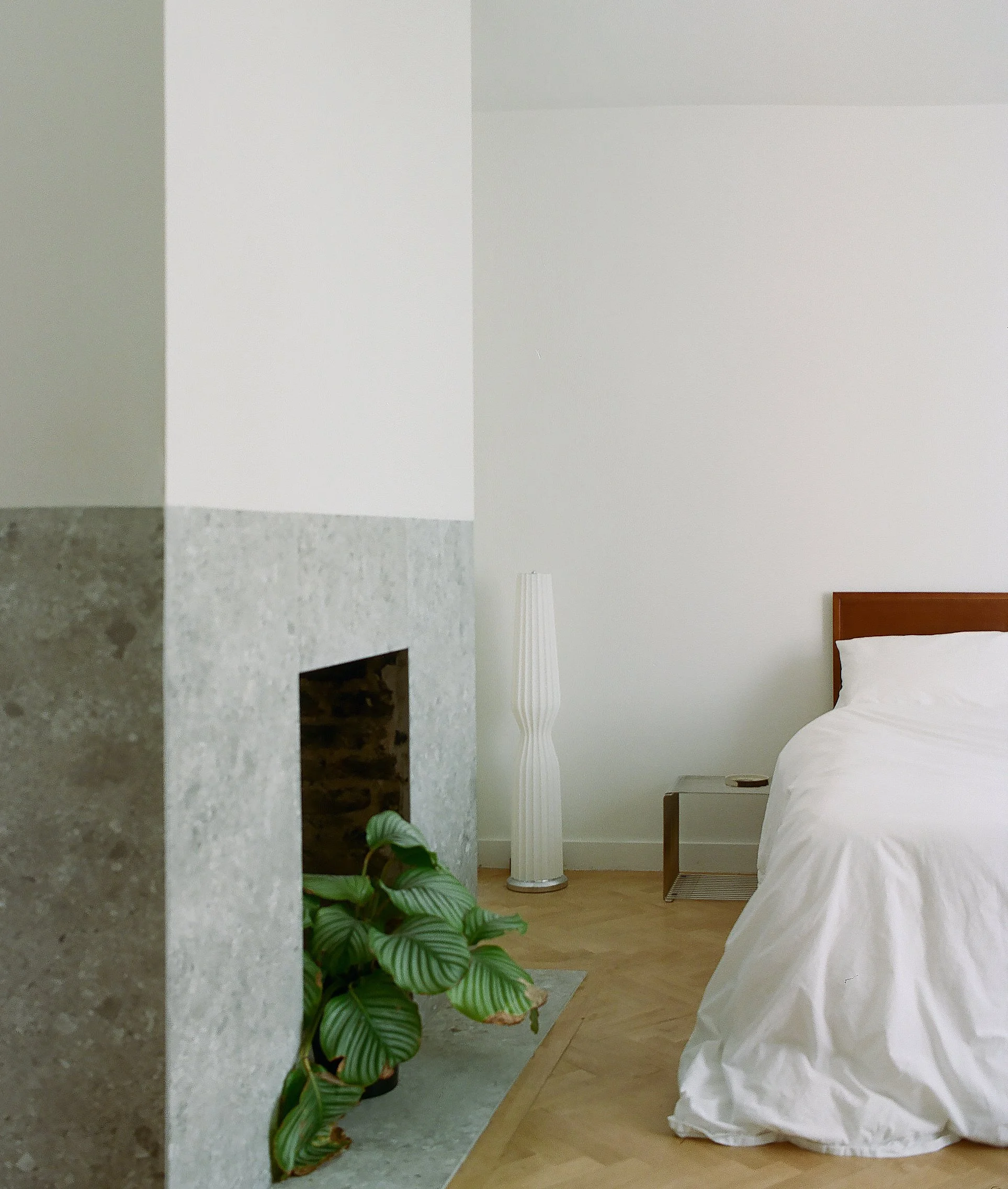Part of a bedroom with a bed covered in white sheets, a wooden headboard, a small side table, a tall white floor lamp, a green potted plant near a fireplace, and light-colored wooden flooring.