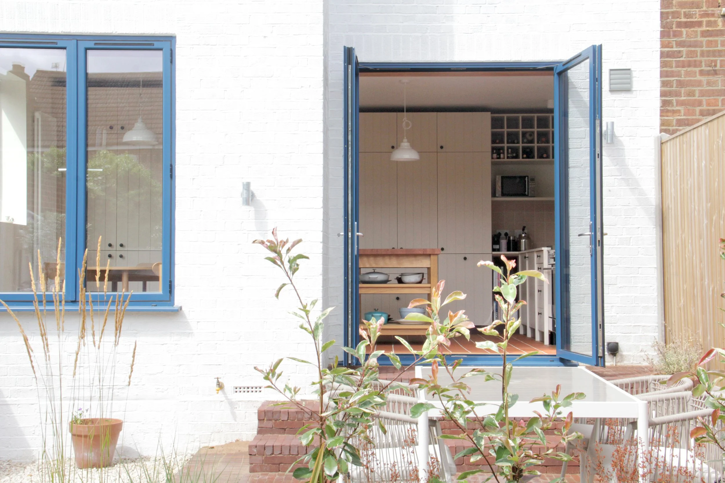 Open glass door leading into a modern kitchen with white cabinets, hanging pendant lights, and various kitchen appliances, outside a white brick house with a window, garden plants, and a patio table.