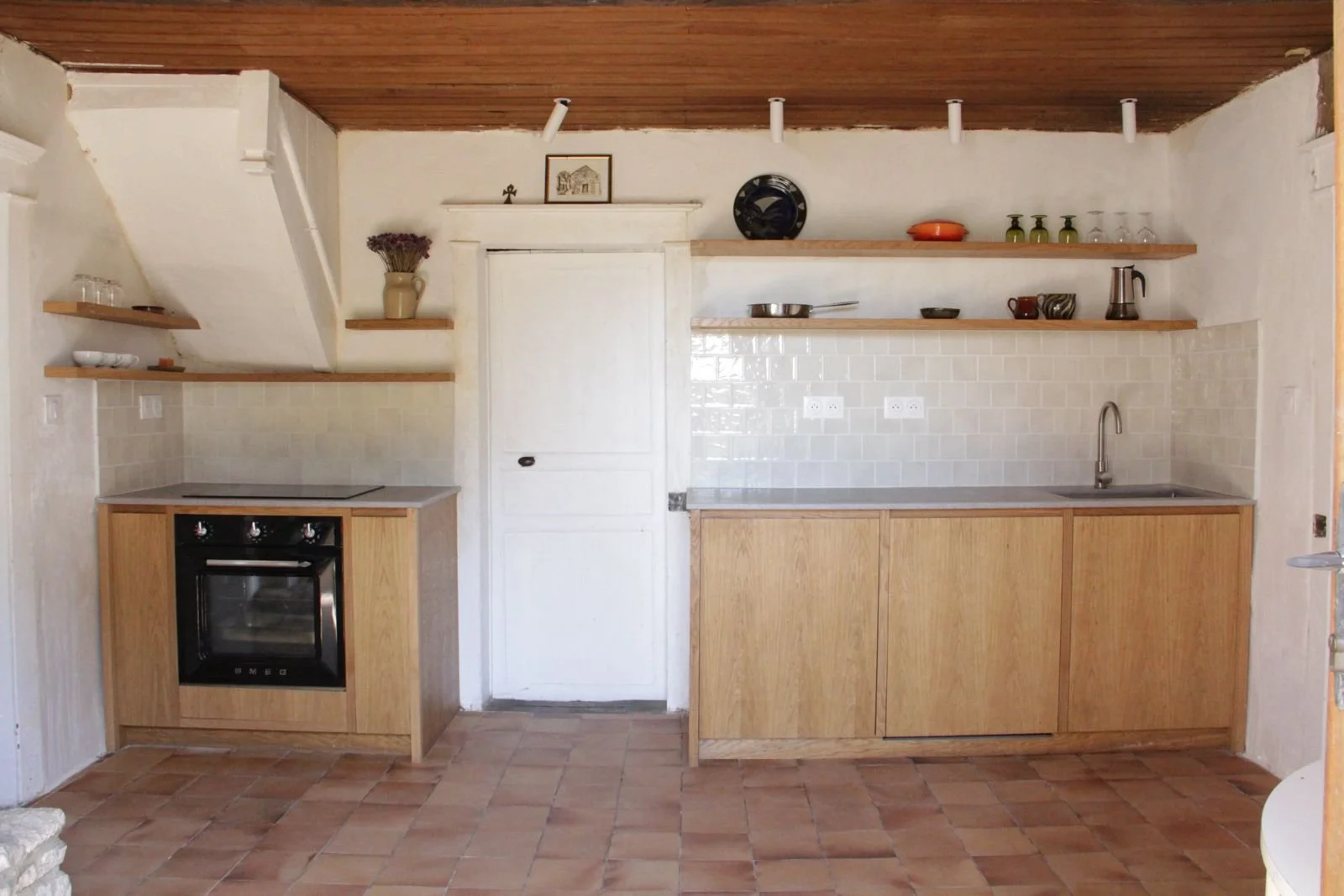 Kitchen with open wooden shelves, a white door, a black oven, a stainless steel sink, and terracotta tile flooring.