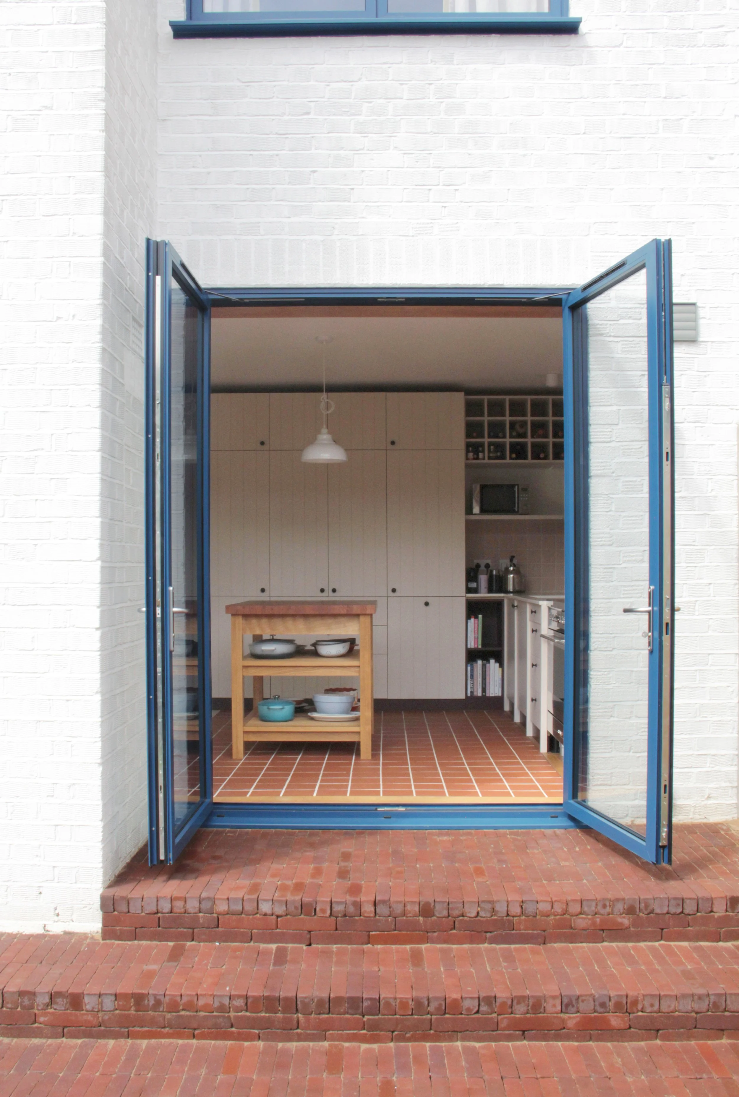 Inside view of a kitchen with white cabinets, a wooden island, and a terracotta tile floor, seen through blue-framed, open double doors on a red brick steps.