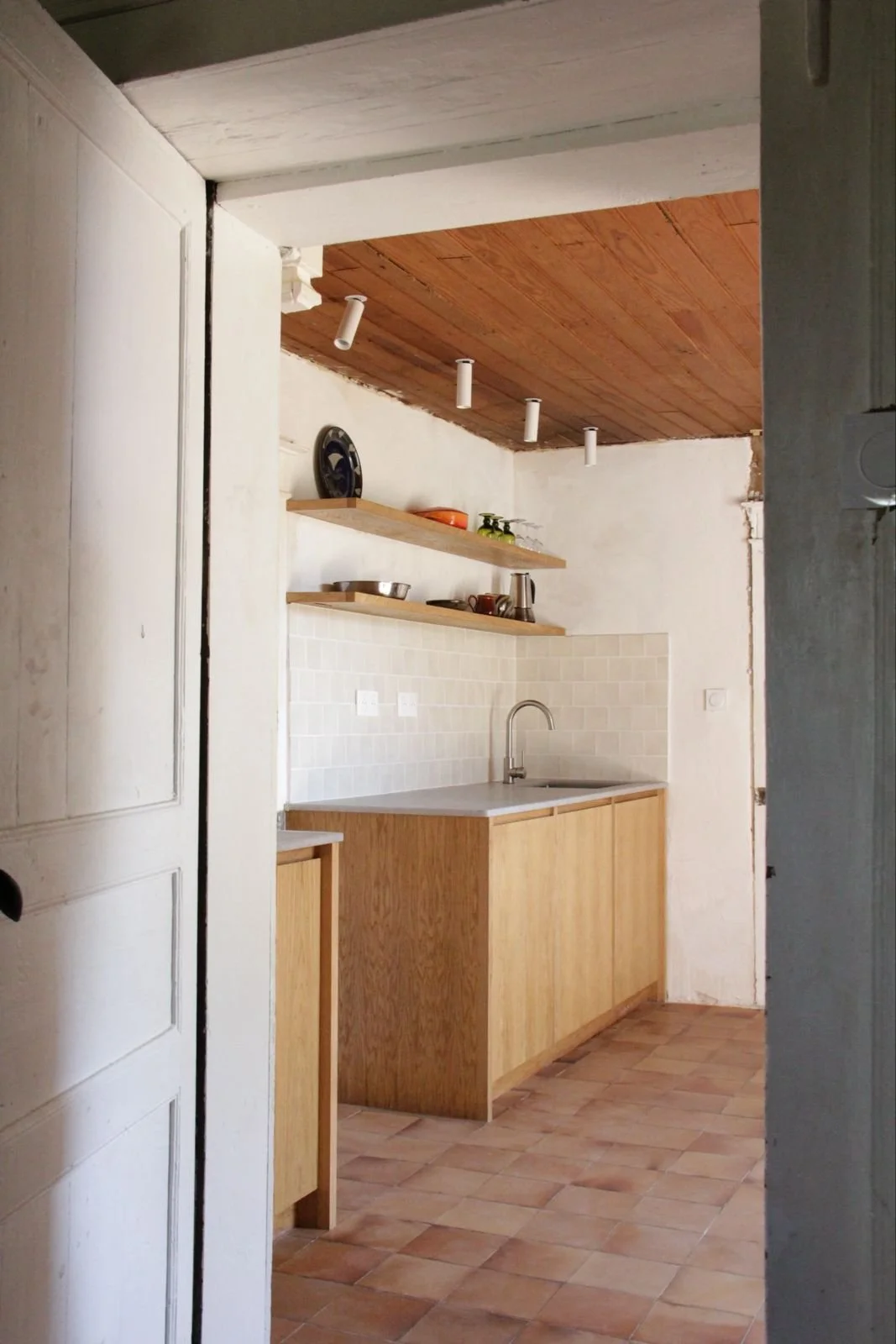 A small kitchen with wooden cabinets, white tiled backsplash, and open wooden shelves holding dishes, glasses, and kitchenware. The ceiling is wood-paneled, and the floor has terracotta tiles.