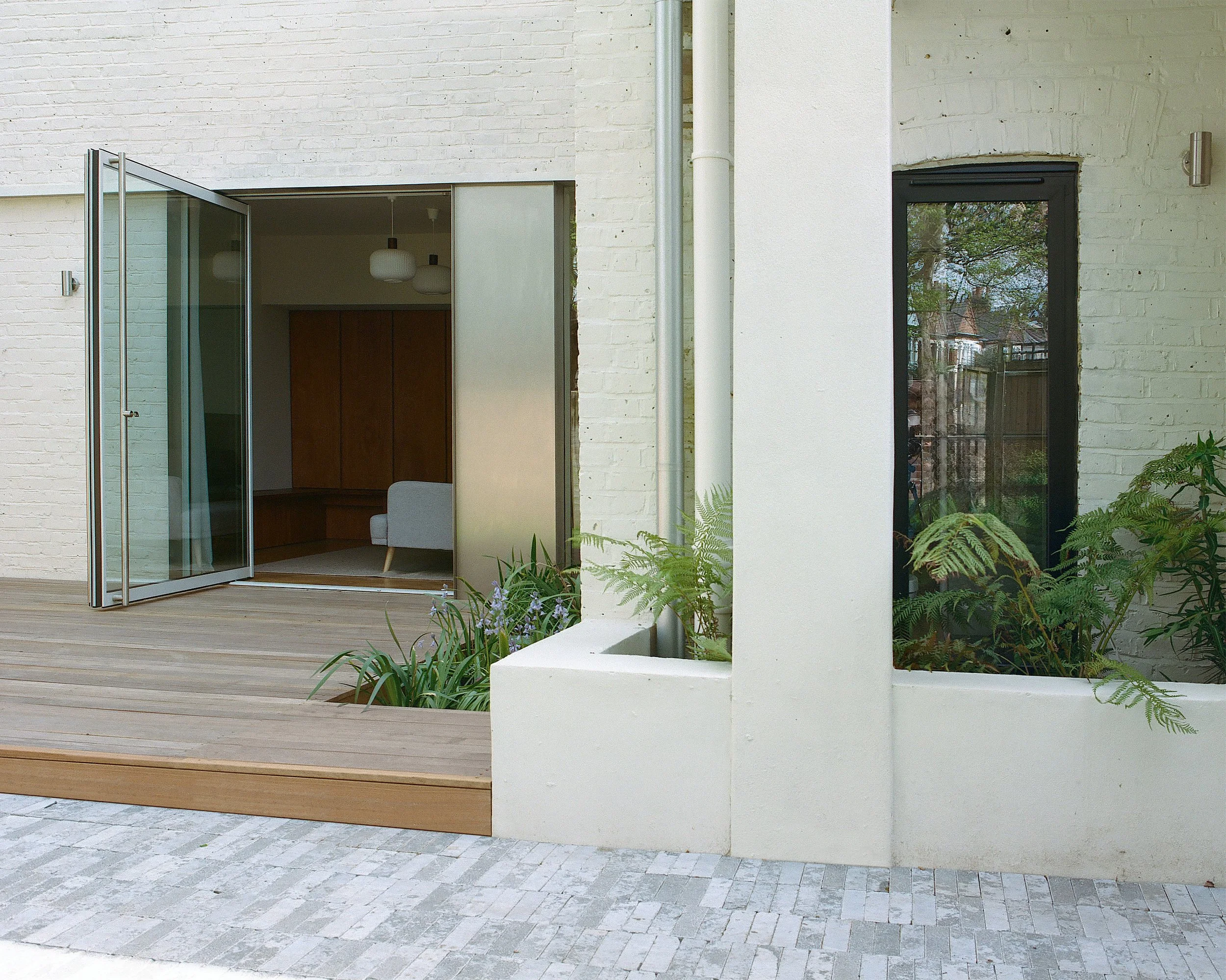 View of a modern house with a wooden deck, an open glass door, and a white brick wall with green plants.