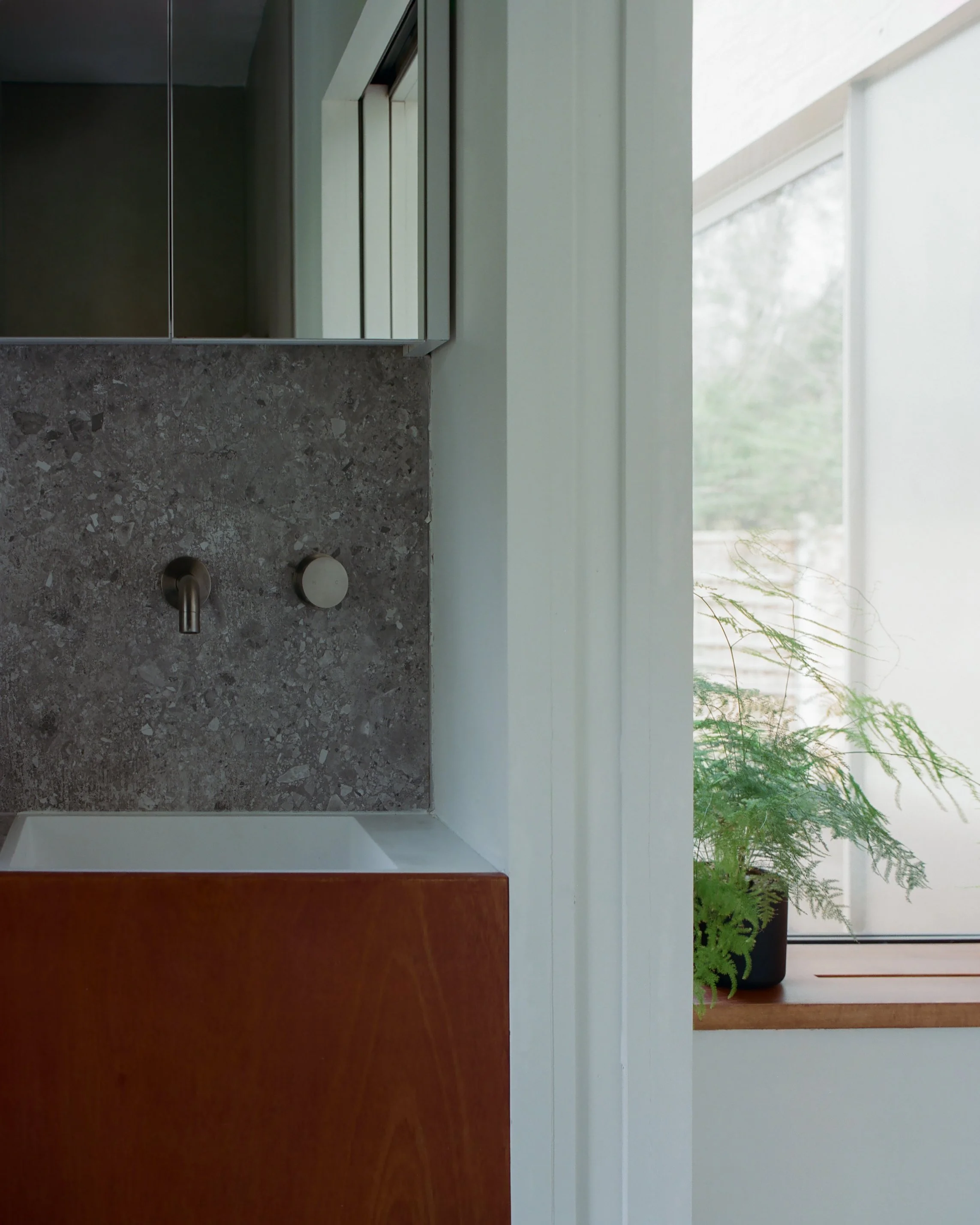 A laundry or utility room with a concrete wall, a faucet, and a wooden cabinet beneath the sink, with a window showing greenery outside.