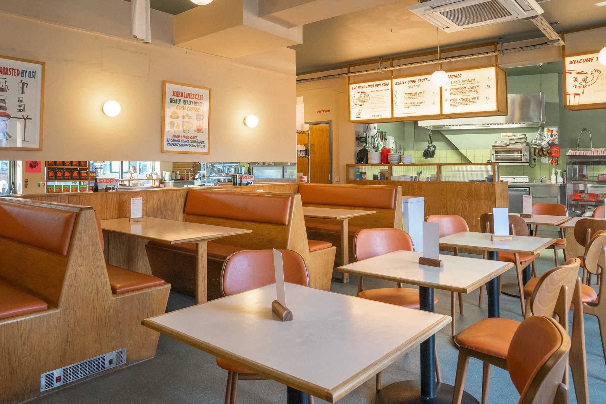 Empty restaurant dining area with wooden tables, orange and wooden chairs, booths, wall art, and a view of the kitchen in the background.