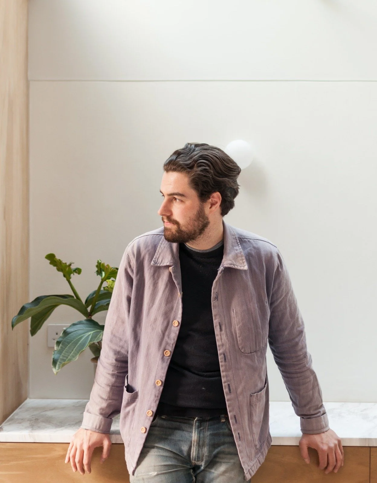 A man with dark hair and a beard leaning against a wooden and marble surface, looking to his left, with a potted plant nearby.